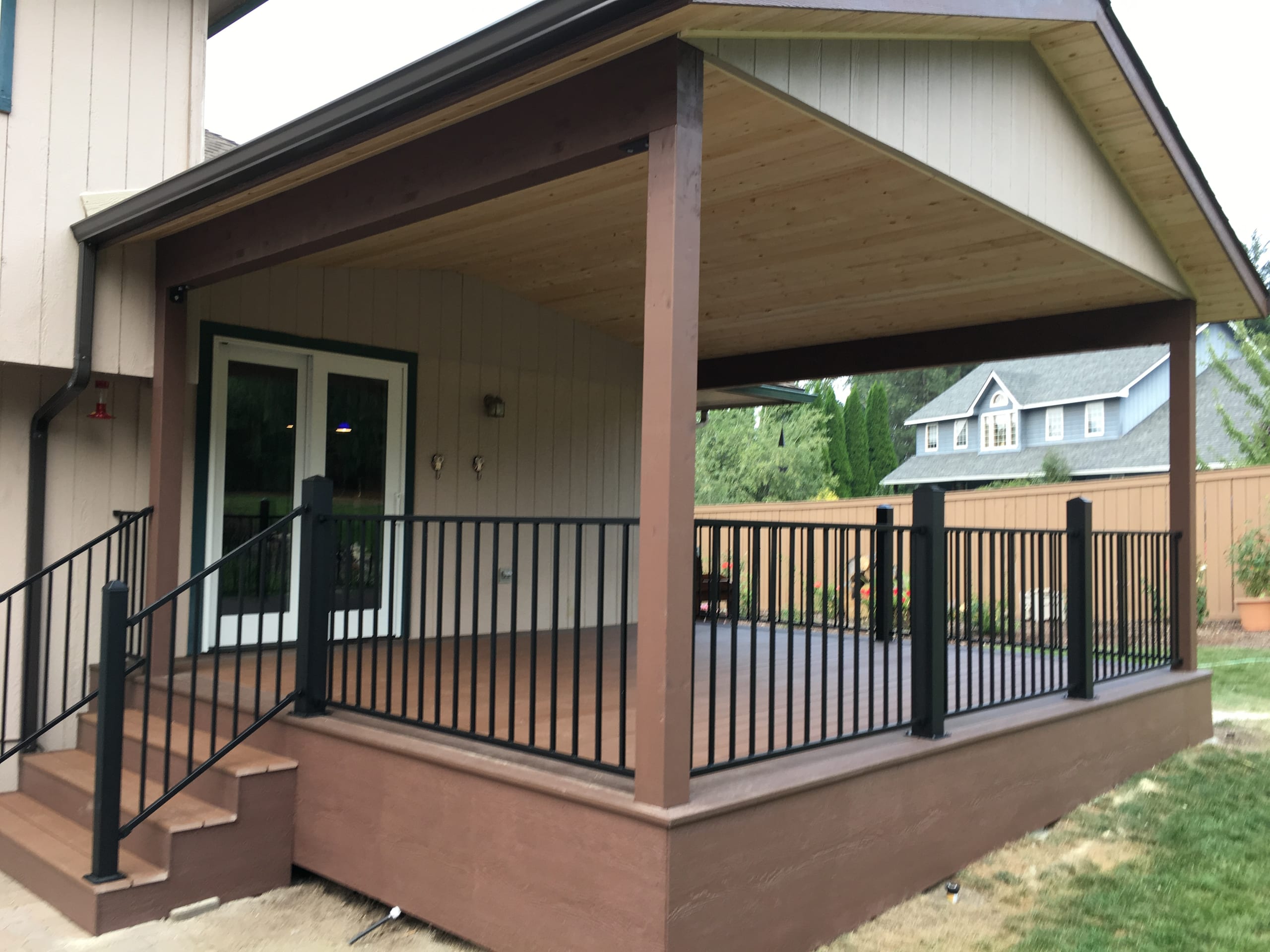 Covered deck with gabled roof, stained wood ceiling, brown posts, black picket railing and stairs