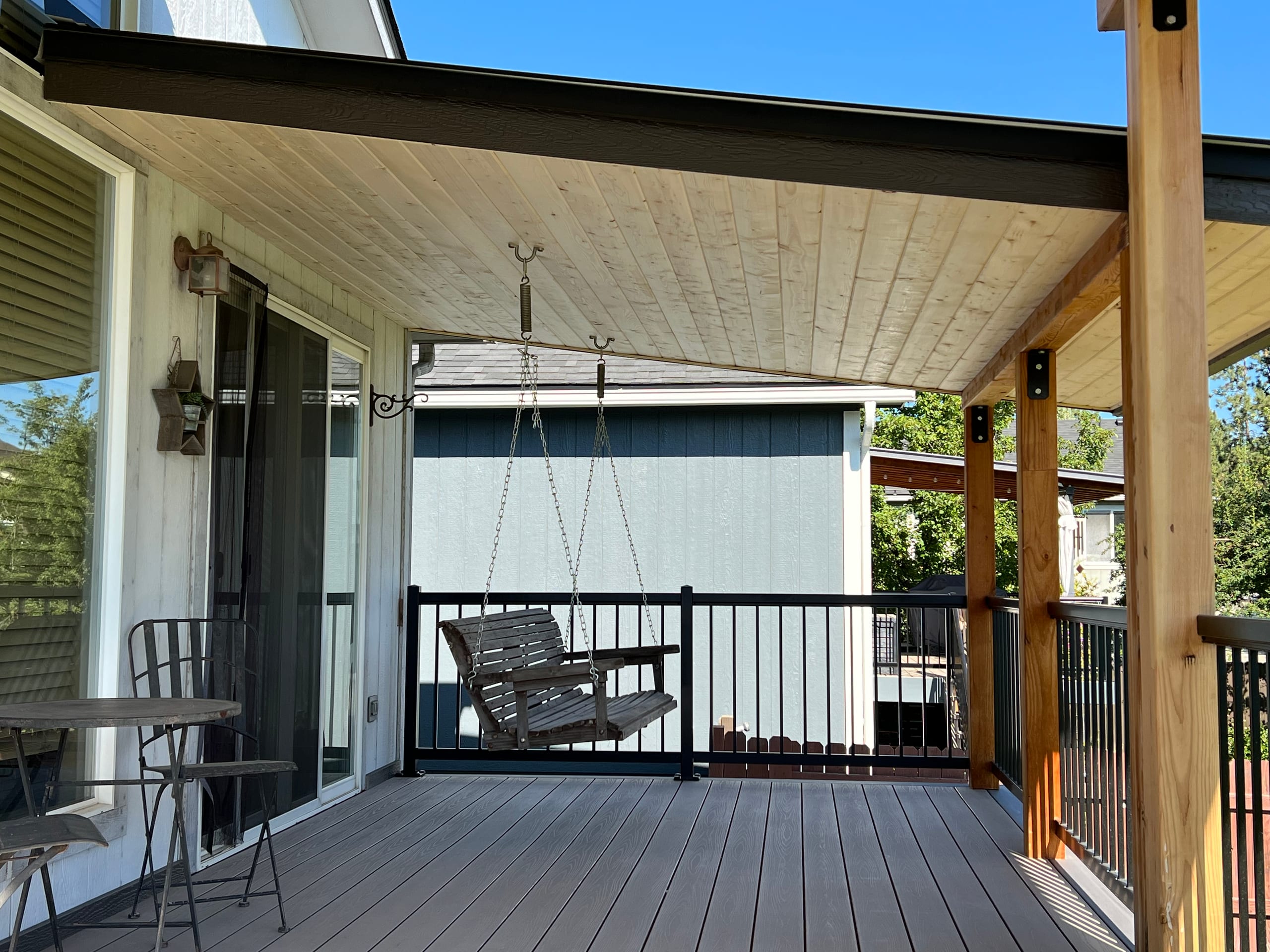 Covered deck with light tongue-and-groove ceiling, cedar posts, porch swing and black railing on grey composite