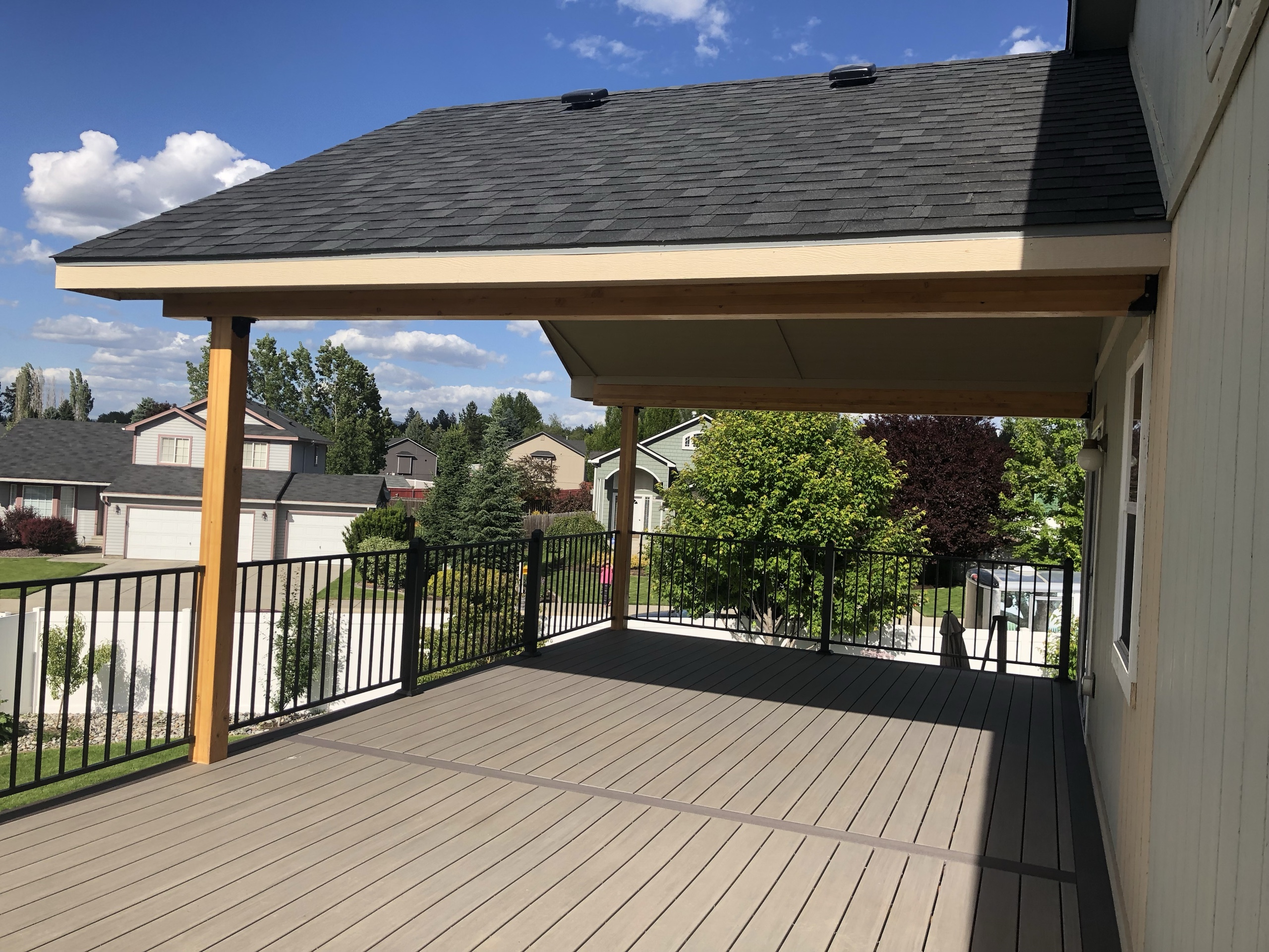 Covered deck with shingle gable roof on cedar posts, brown composite decking, black railing, neighborhood view