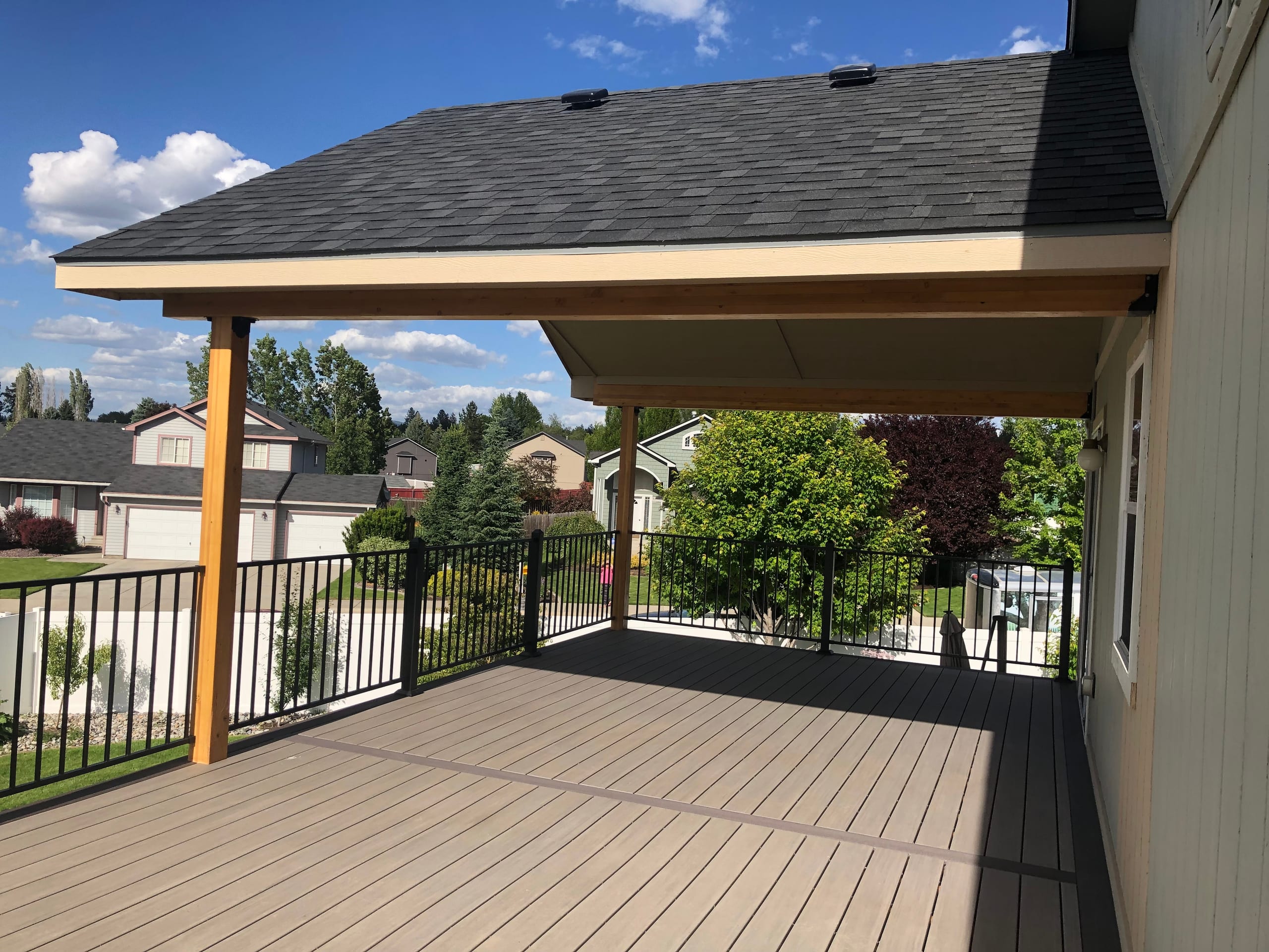 Covered deck with shingle gable roof on cedar posts, brown composite decking, black railing, neighborhood view