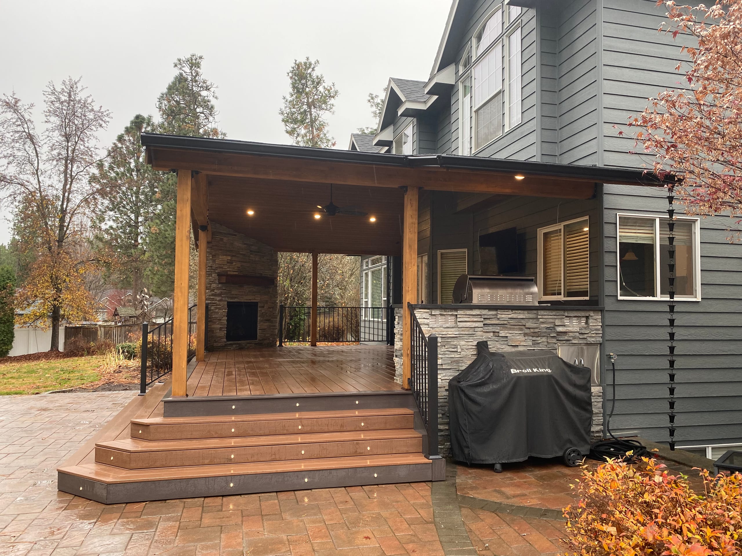 Covered deck with stone fireplace, outdoor kitchen, stained timbers and wide stepped paver landing, grill under cover