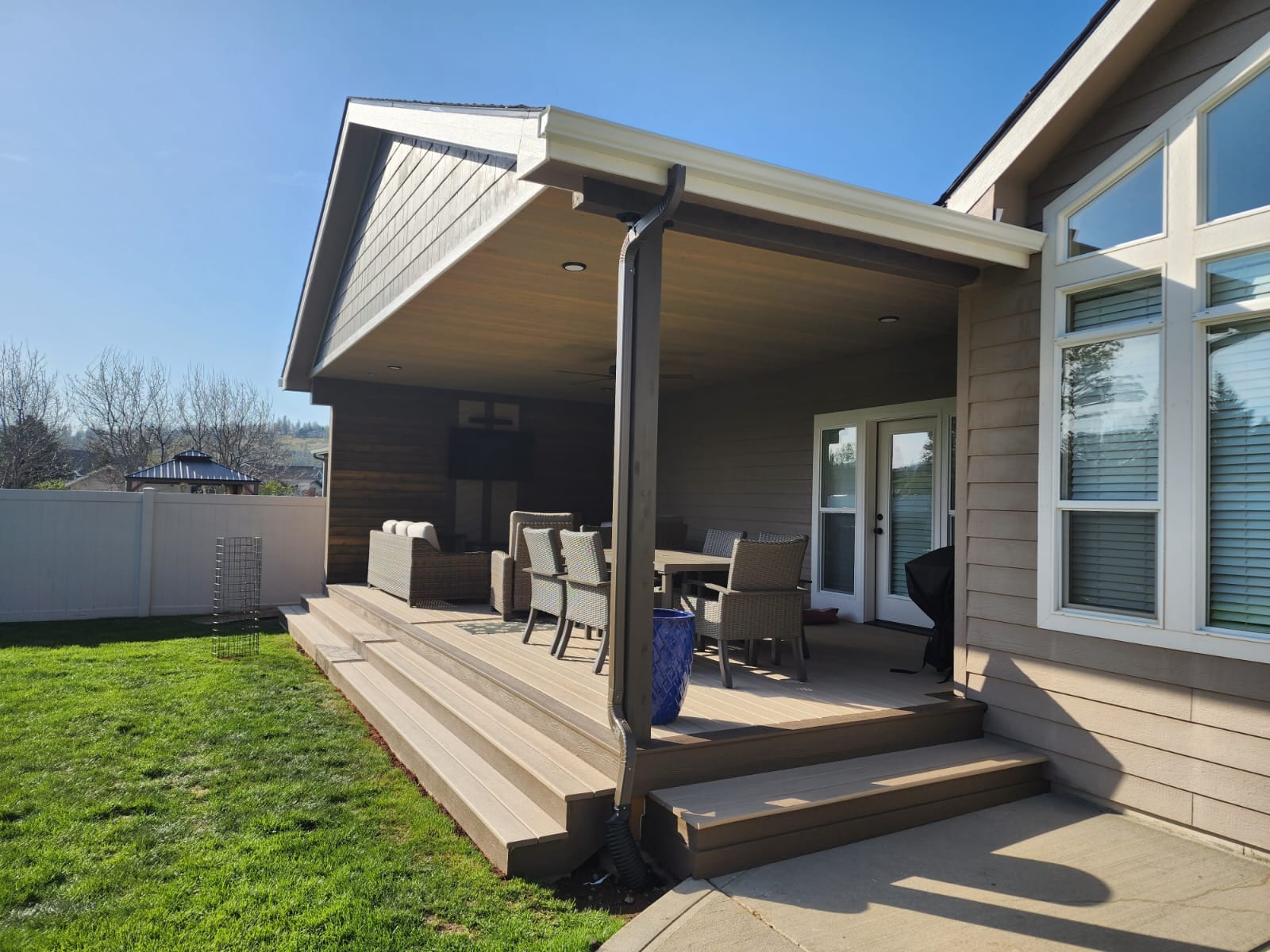 Covered deck with tan composite wide steps, outdoor furniture, gabled roof and green lawn under blue sky
