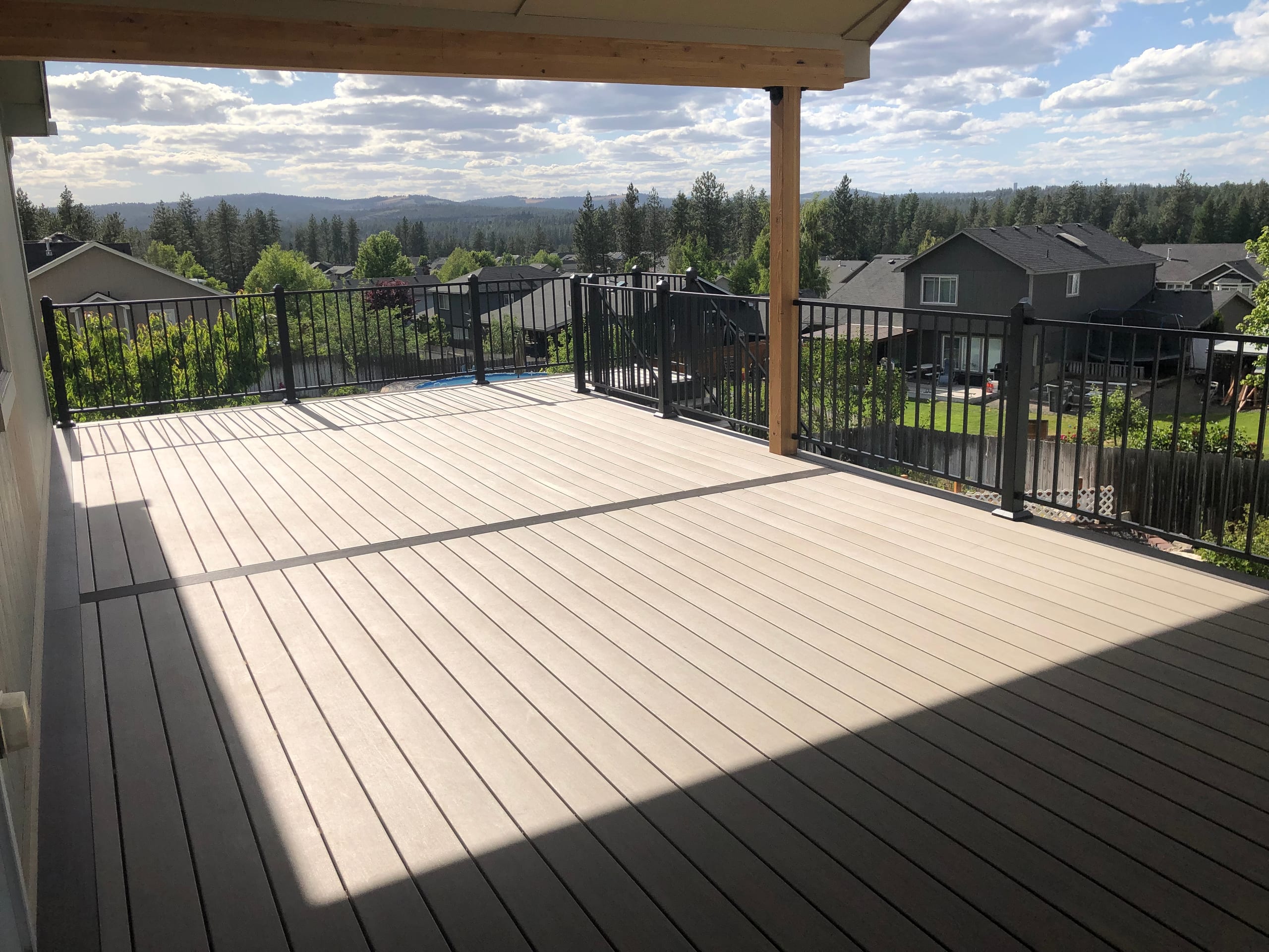 Covered deck with tan decking and black railing overlooking a neighborhood and wooded hills