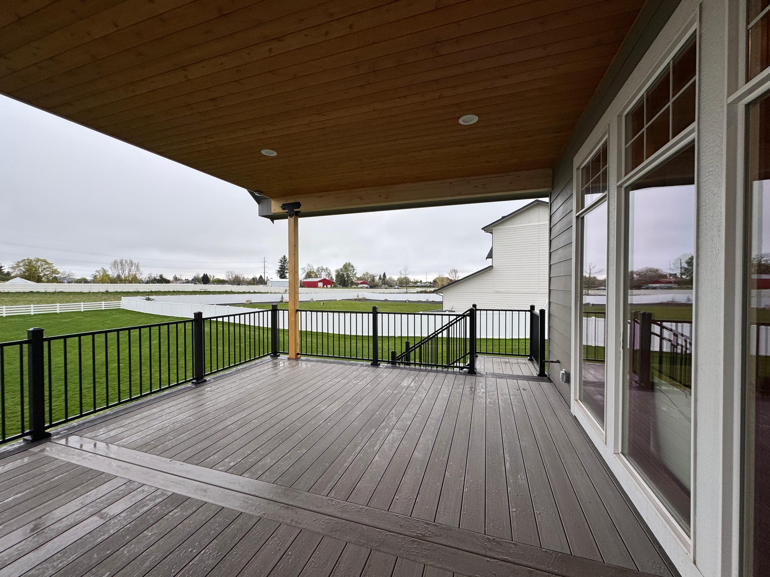 Covered deck with wood ceiling, glass doors and black metal picket railing looking across white-fenced pasture
