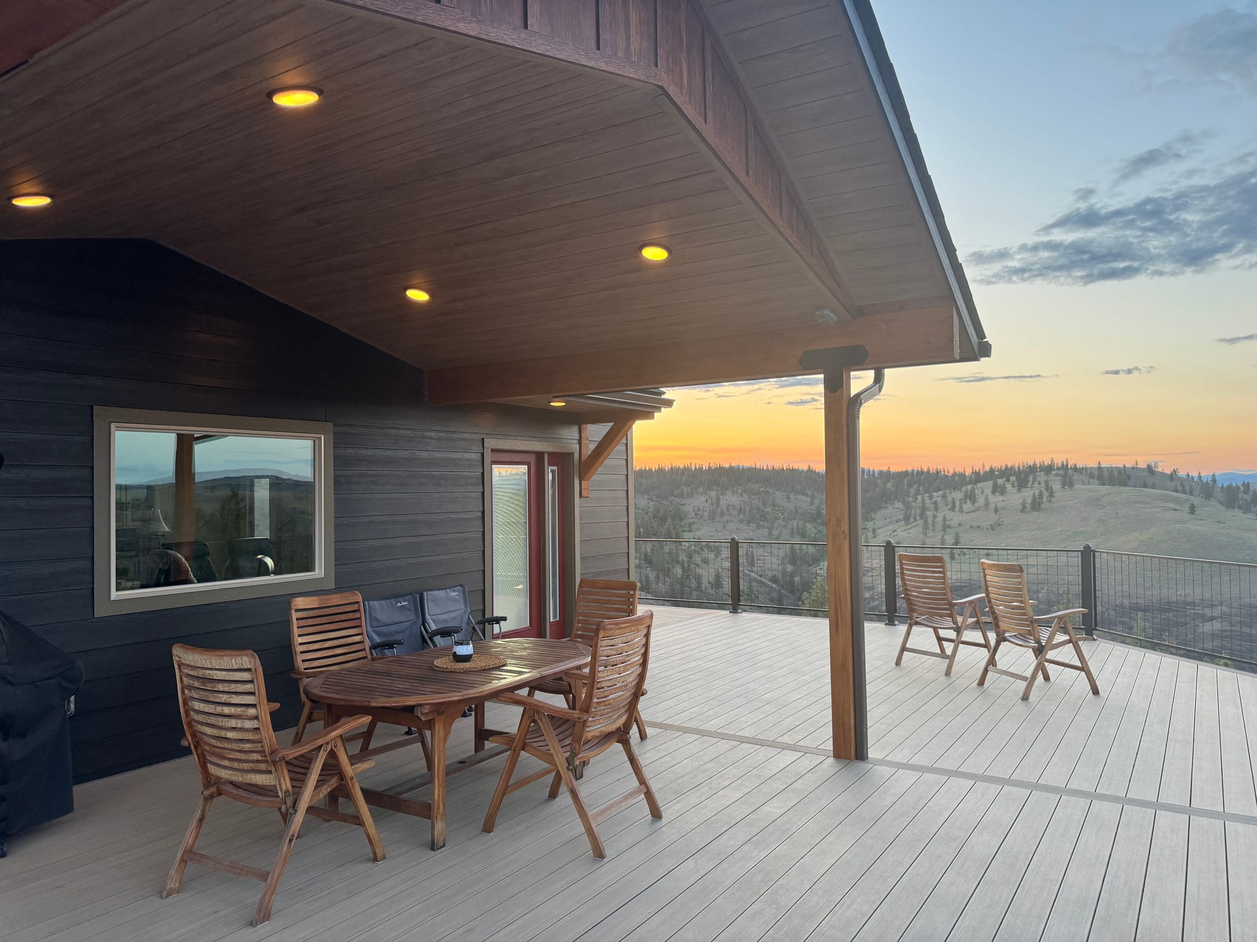 Covered gray composite deck at sunset with teak dining set, cedar ceiling lights and hills view