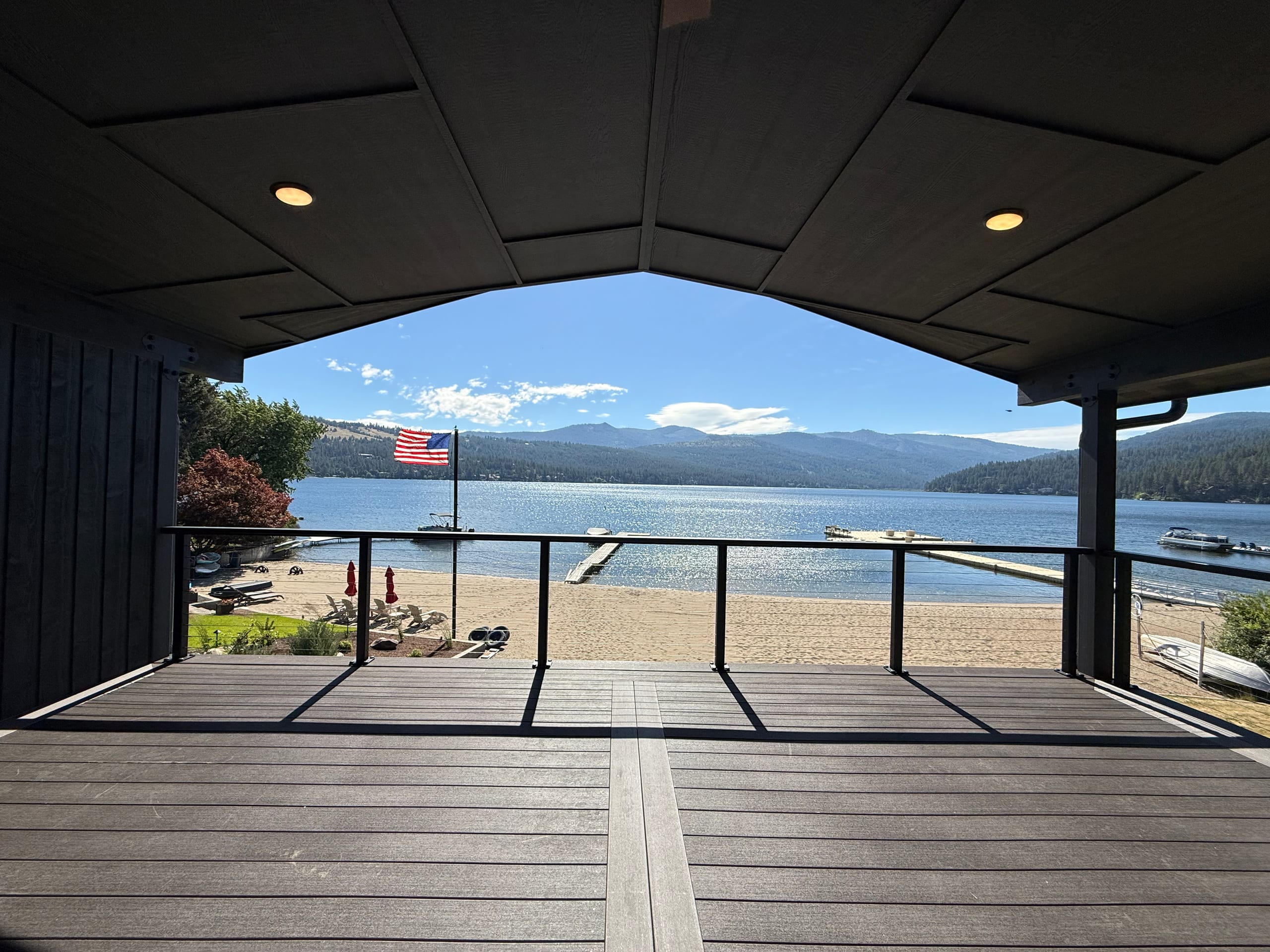 Covered lakefront deck looking out through gable to beach, dock, flag and mountains, dark composite boards and cable railing