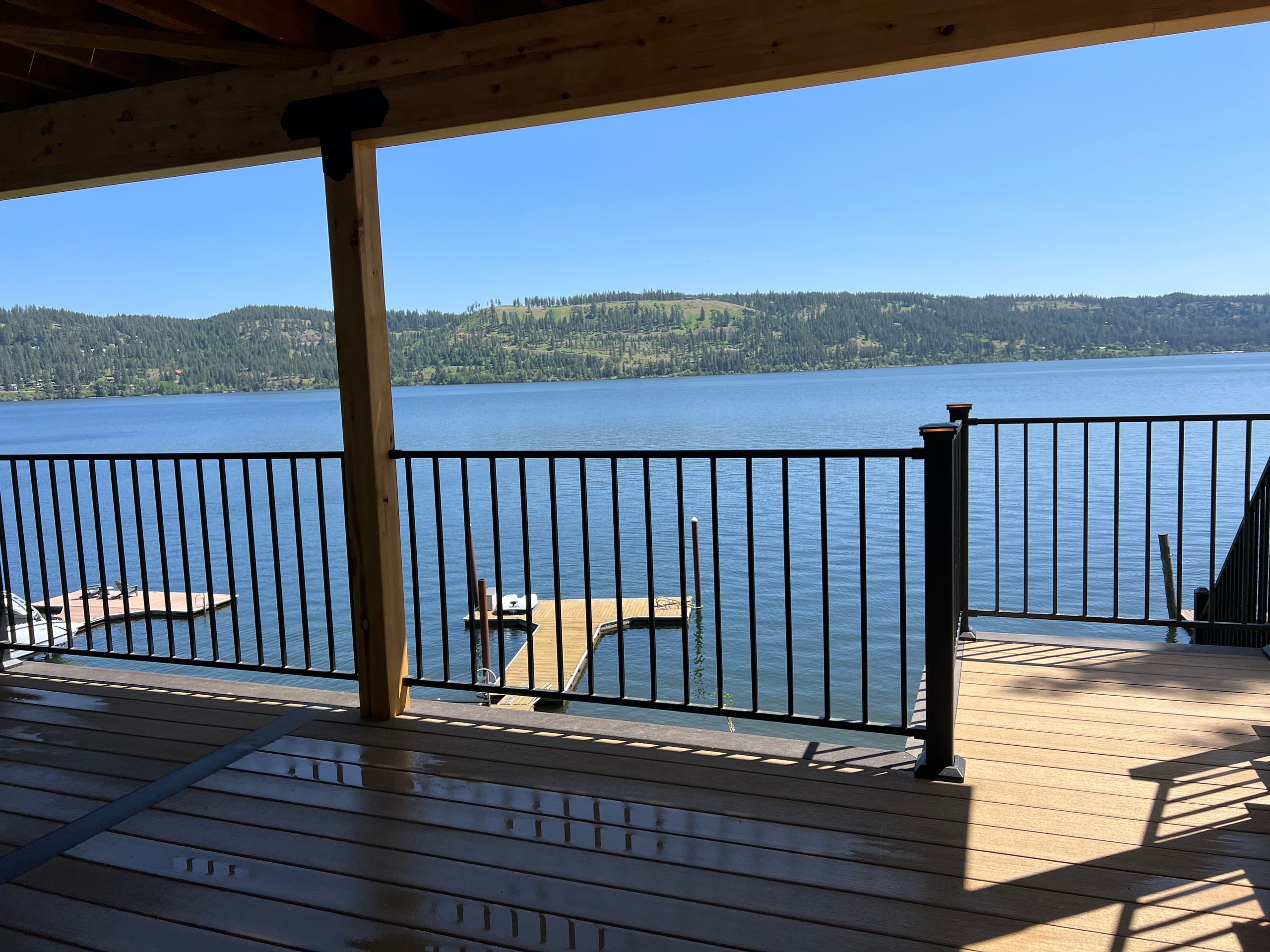 Covered lakeside deck with black picket railing framing a wide blue lake view, dock below and sunny shadows