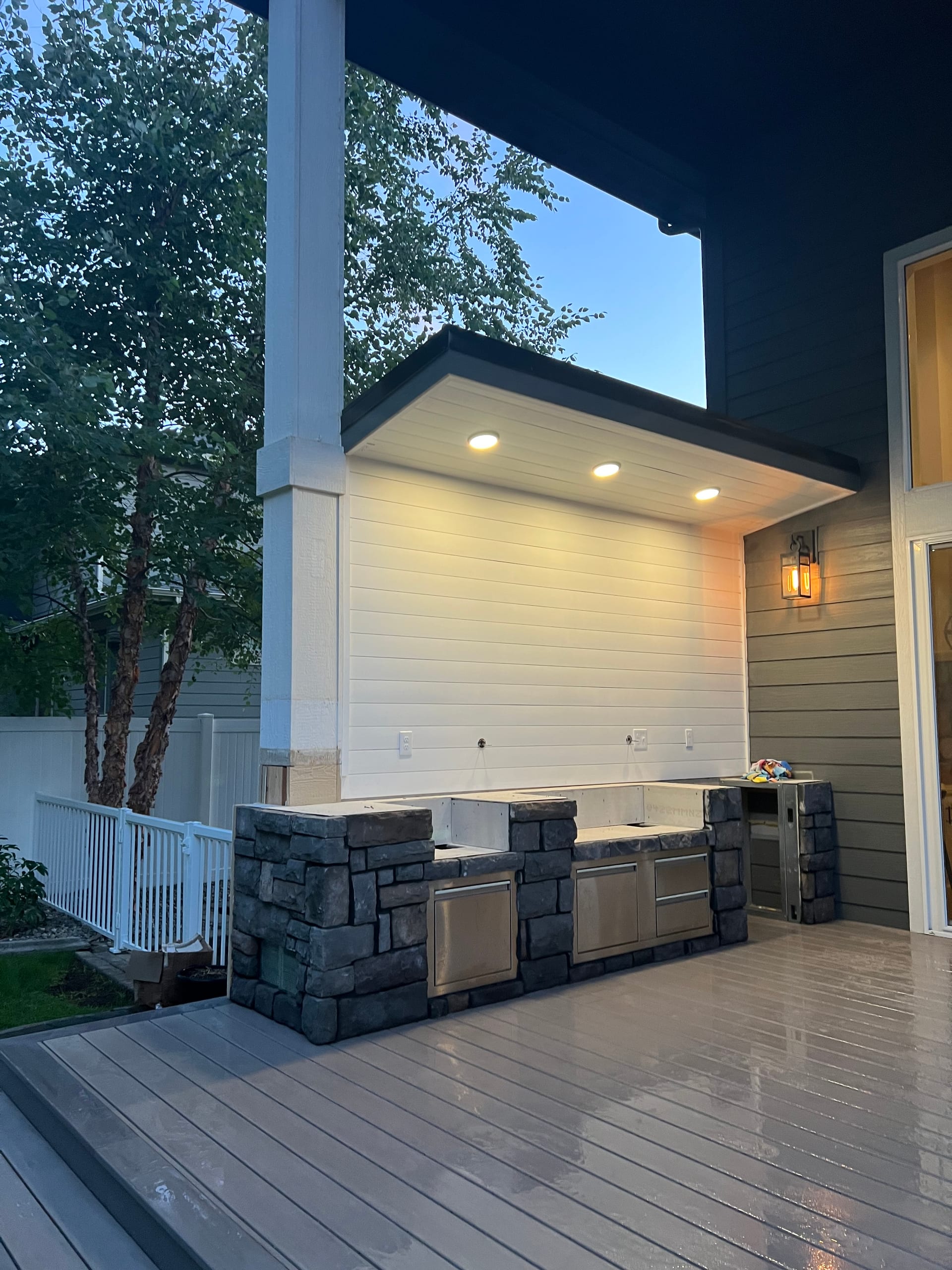Covered patio kitchen with stone-clad counters and stainless appliances under recessed lights, wet gray composite deck