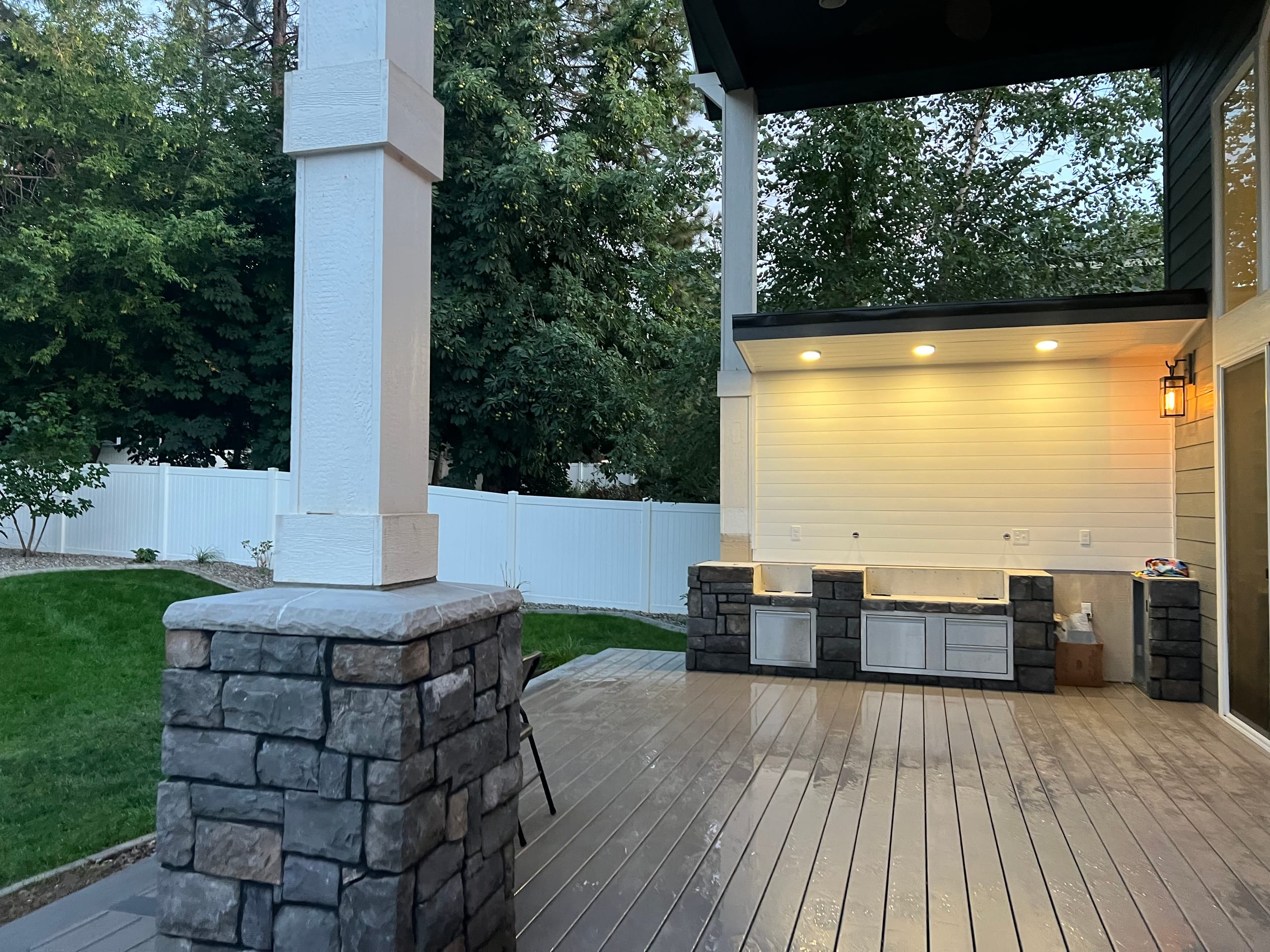Covered patio with stone-based columns, built-in outdoor kitchen under recessed lights and wet gray composite deck