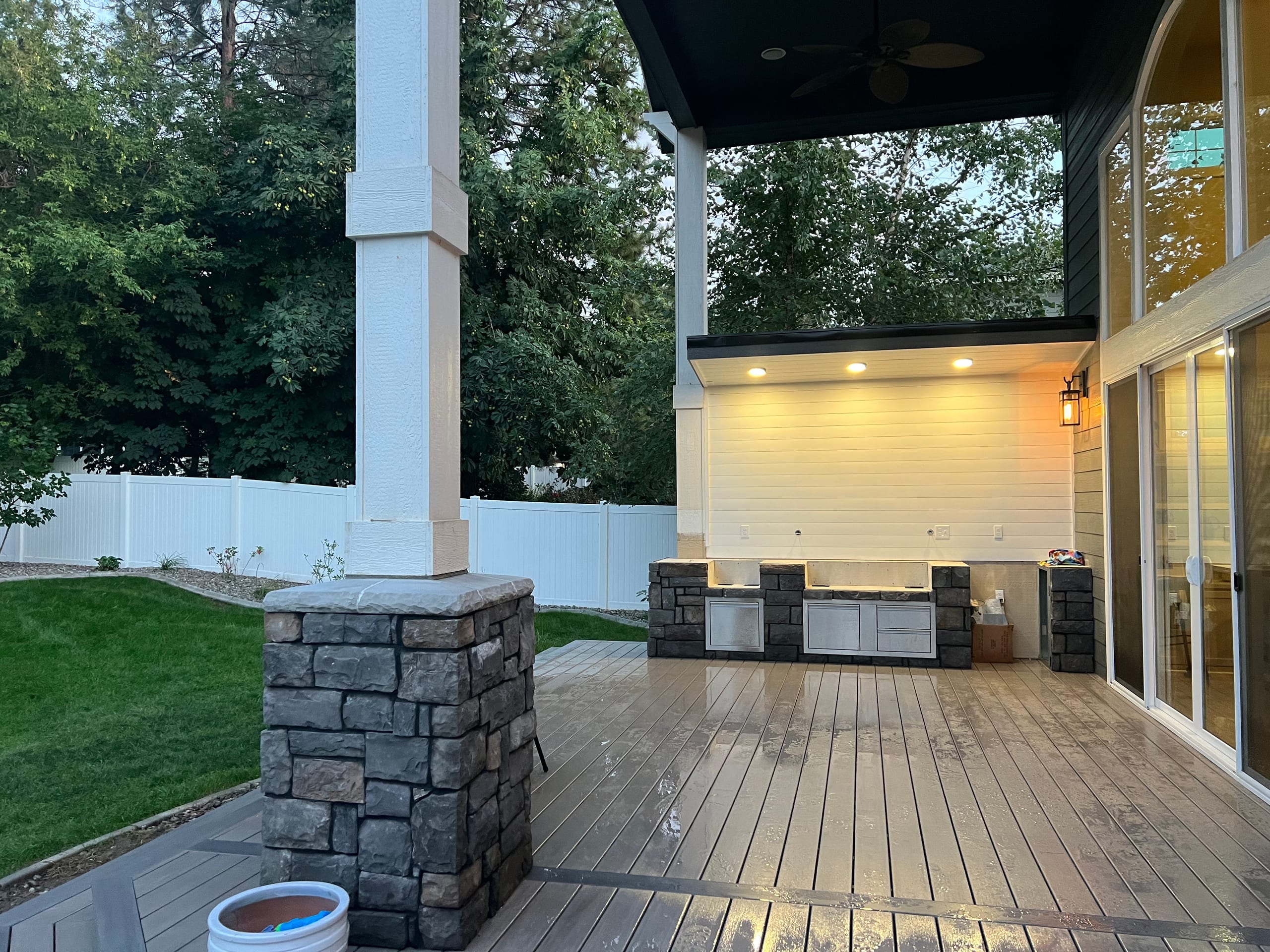 Covered patio with stone-and-white columns, dark ceiling, outdoor kitchen with lit wall and wet gray composite deck