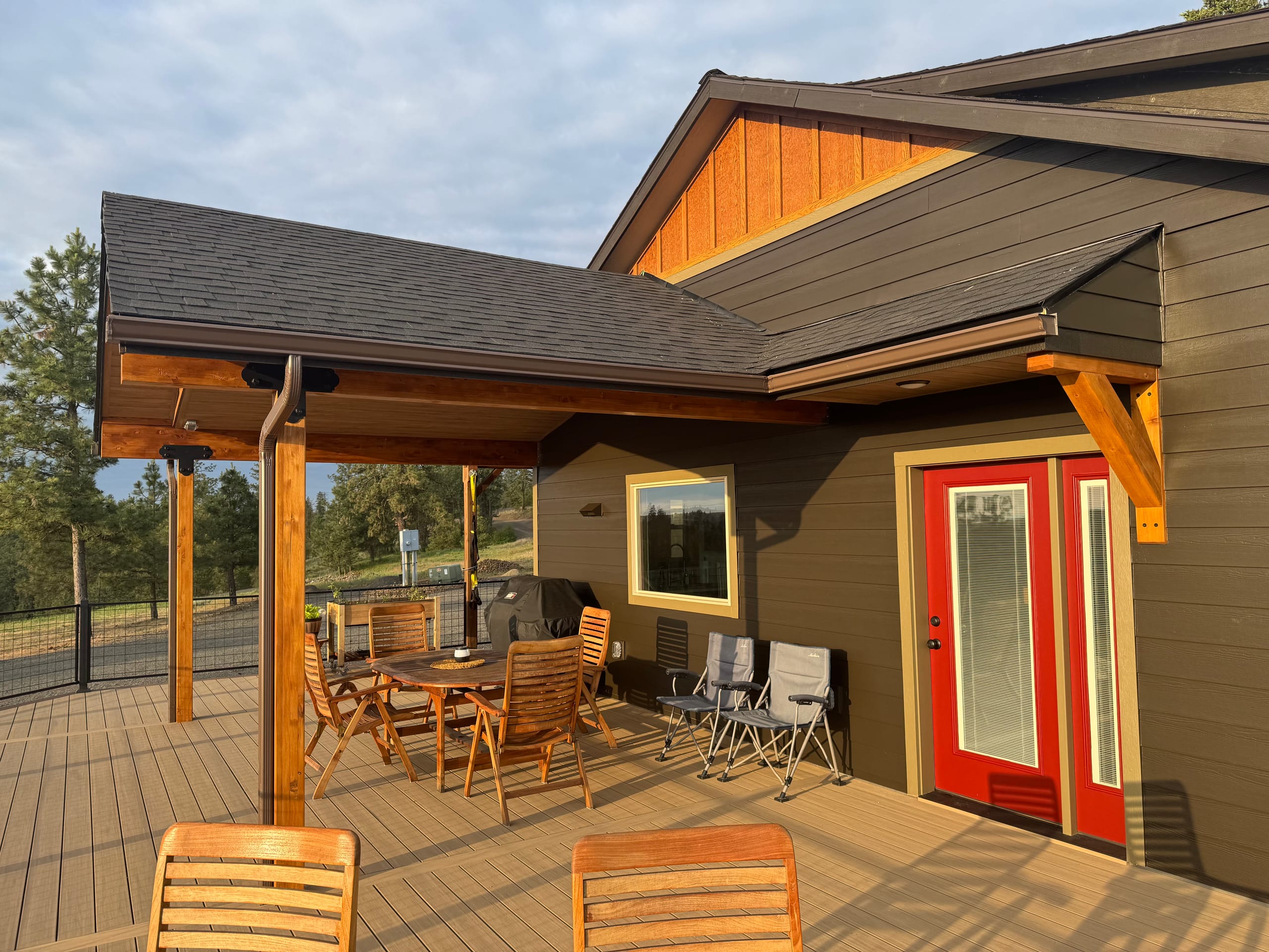 Covered patio-style deck under shingled roof with cedar beams, patio table, wood chairs and red doors on brown house