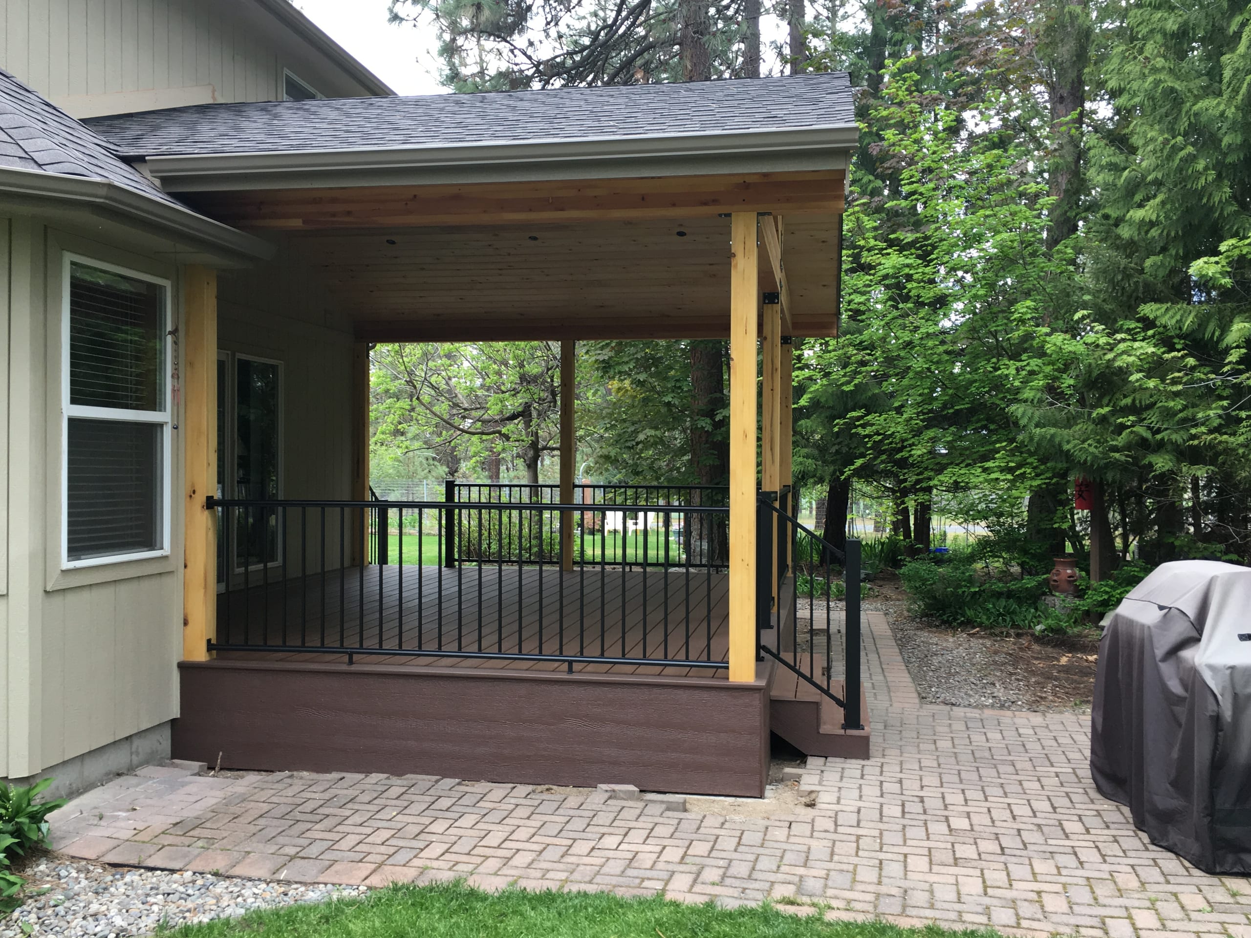 Covered porch addition with dark brown composite deck, cedar posts, black railing and paver patio, green house