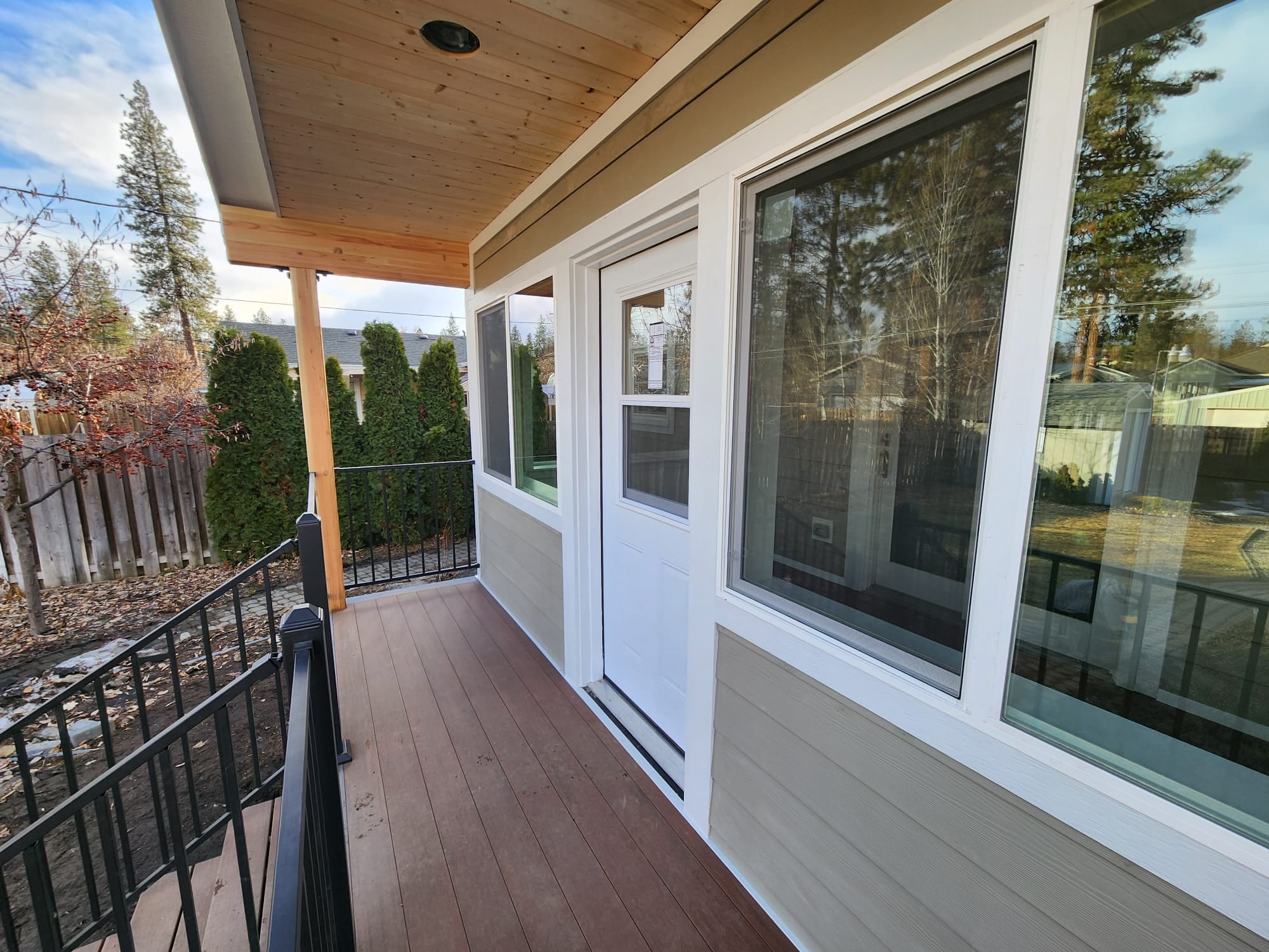 Along the covered porch with brown composite deck, white door, large windows and black picket railing, stained wood ceiling