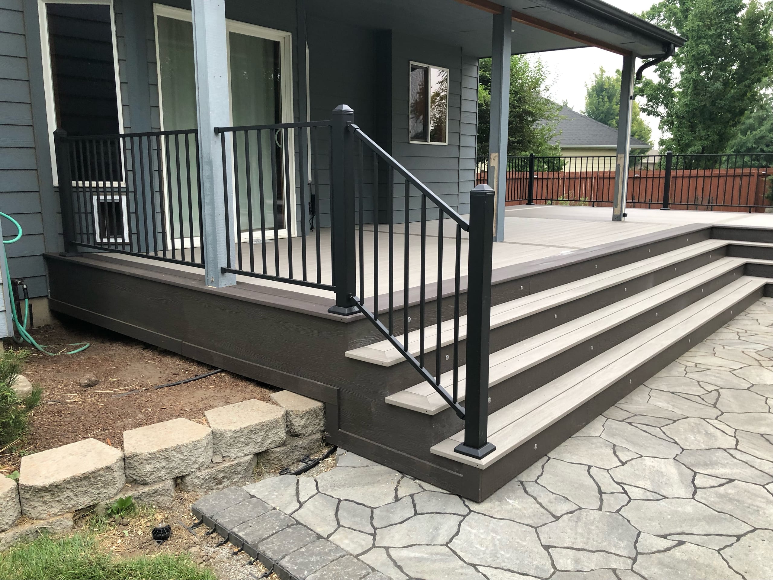 Covered porch deck with grey composite surface, dark fascia and black metal railing, wide steps onto a flagstone patio