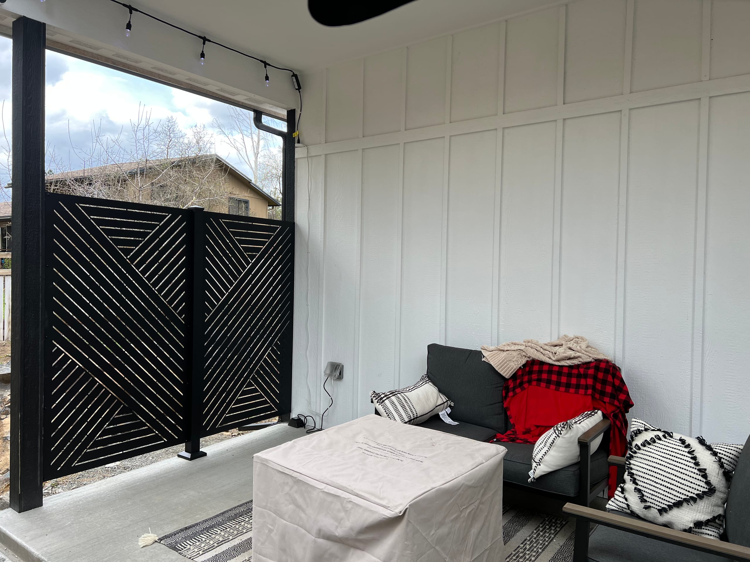 Covered porch with white board-and-batten wall and black laser-cut privacy screen panel, patio furniture and string lights