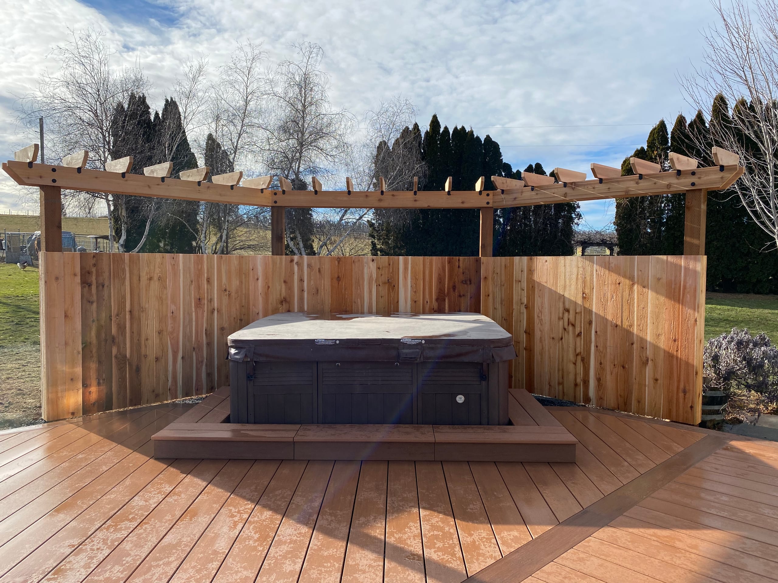 Curved reddish composite deck with hot tub sunk in a platform, cedar privacy fence and curved pergola behind, field view