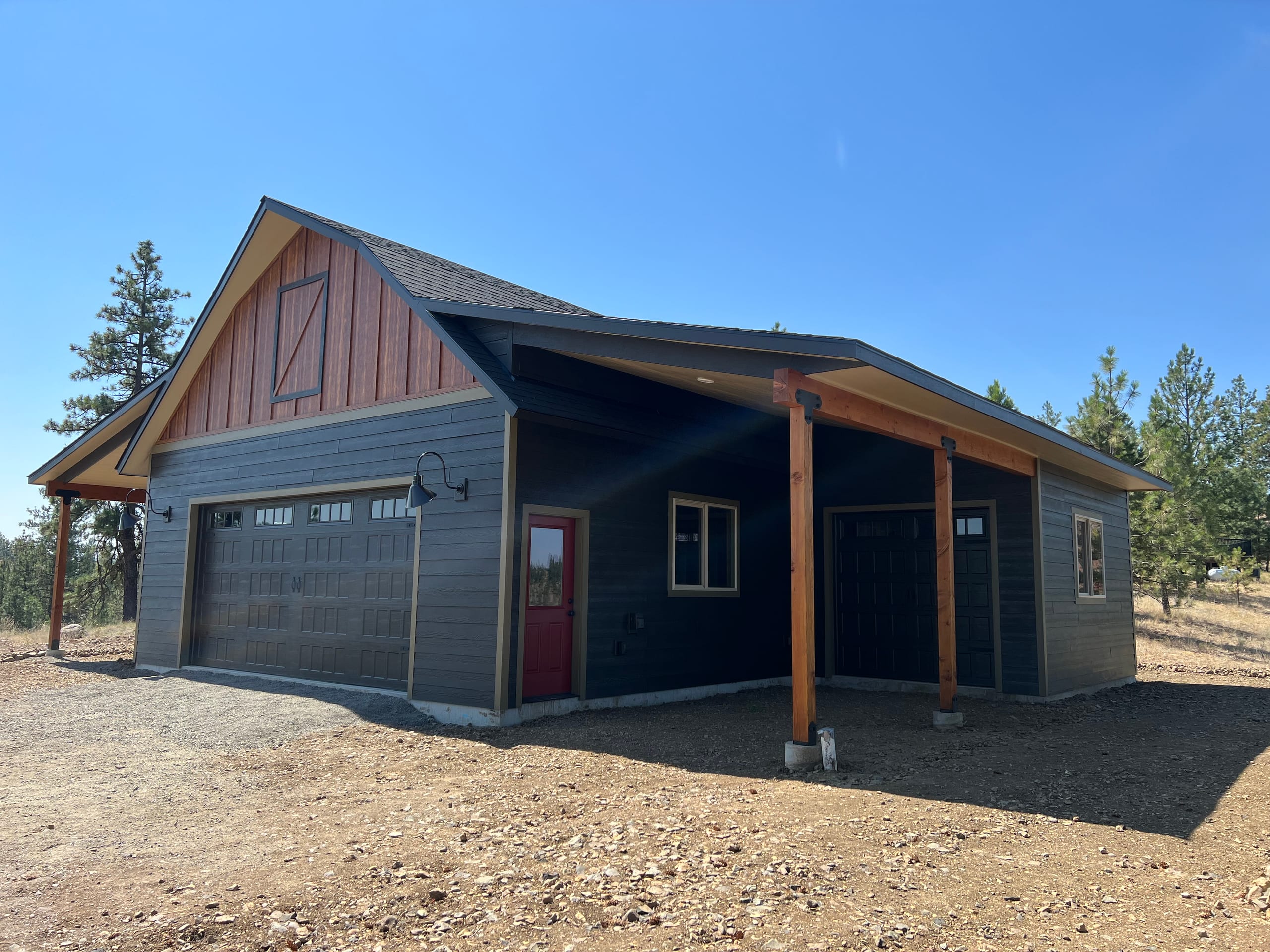 Dark barn-style garage with cedar board-and-batten gable, red door and covered side porch on bare dirt, clear sky