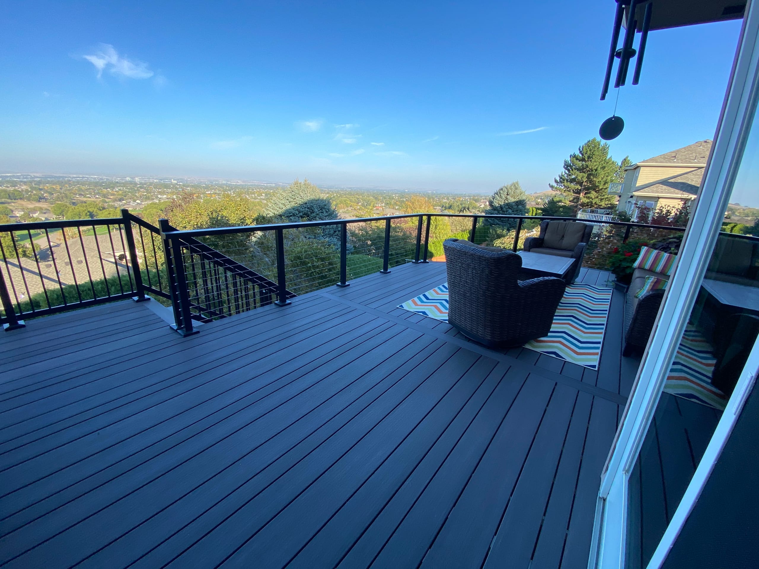 Dark blue-grey composite deck with black railing, wicker seating and city/valley view under blue sky