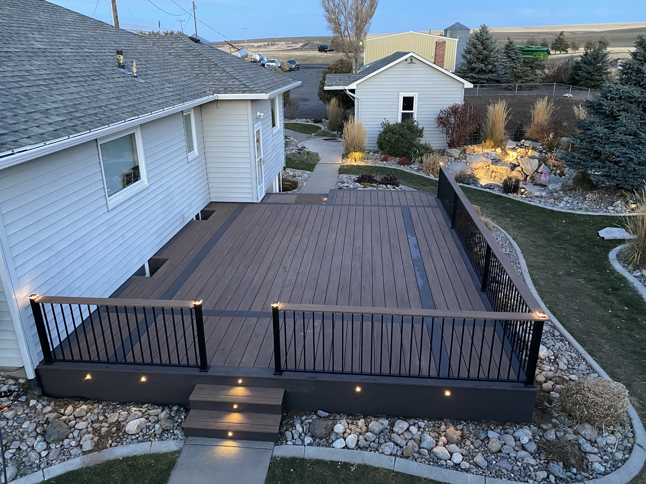 Dark brown composite deck with accent stripe, black railing and lit fascia and steps at dusk, landscaped rock border