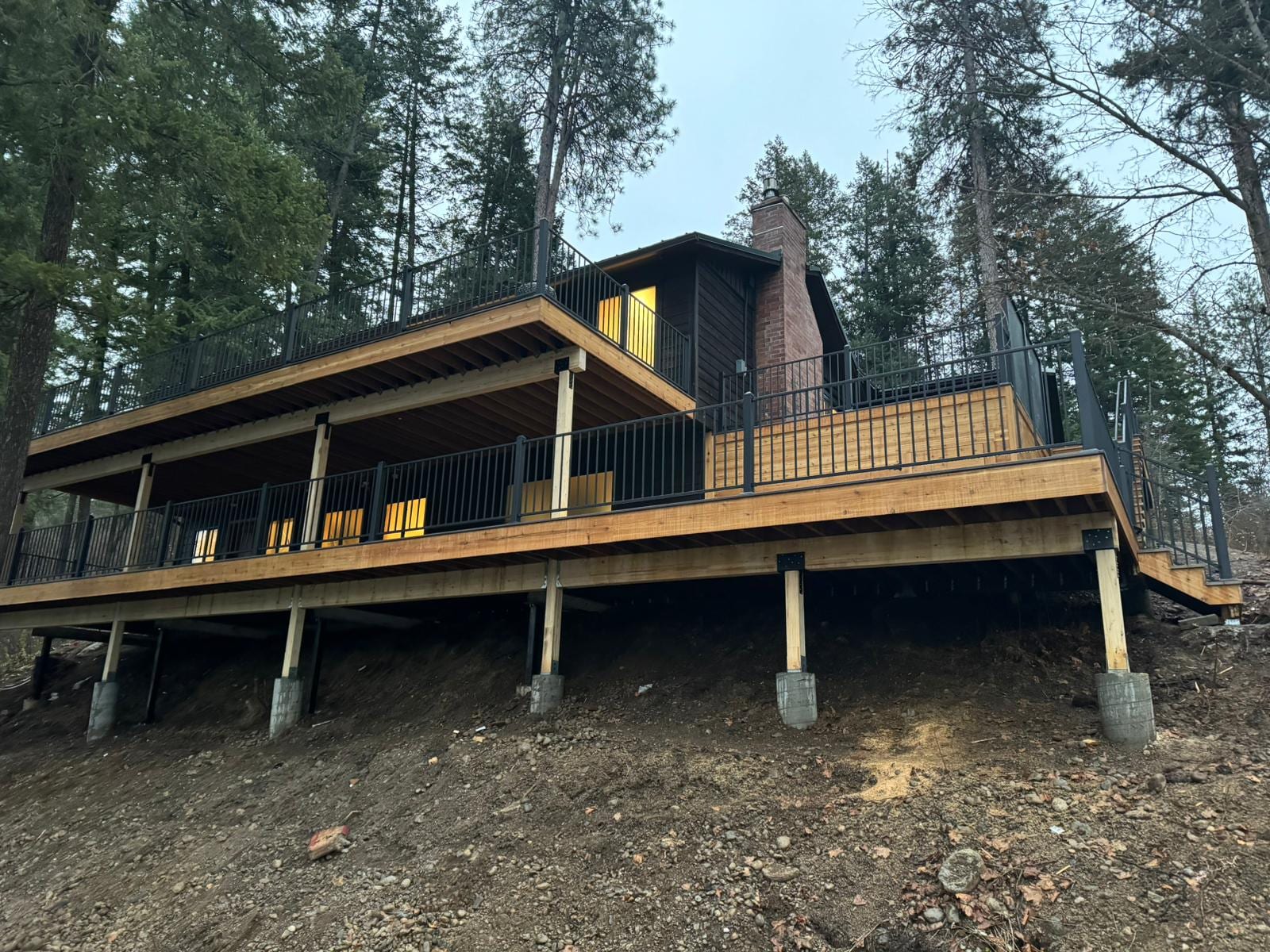 Dark cabin on a wooded slope with large elevated cedar-look deck on posts, black metal railing and covered section