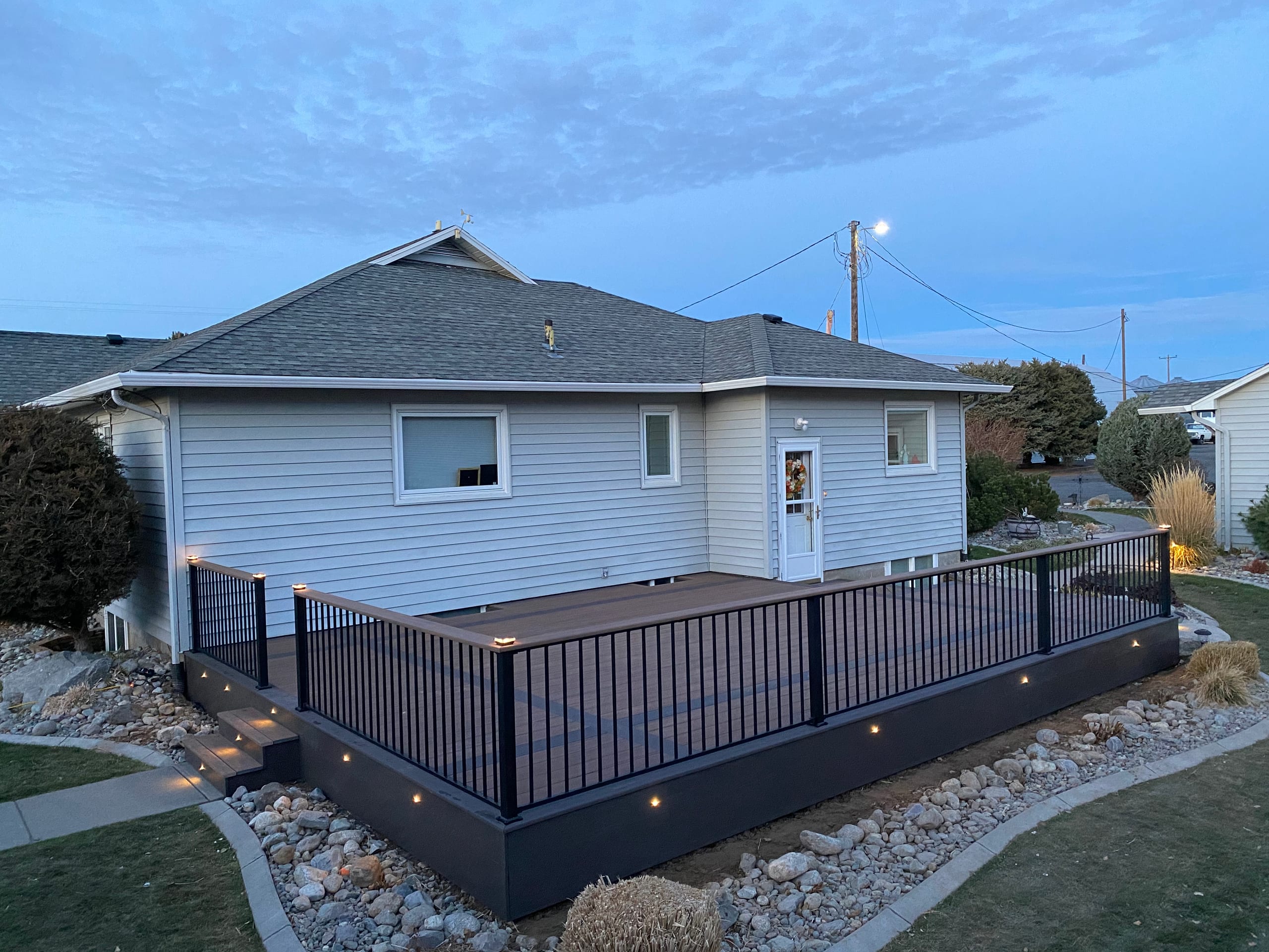 Dark composite deck with black picket railing, lit fascia and steps at twilight, rock border and lawn