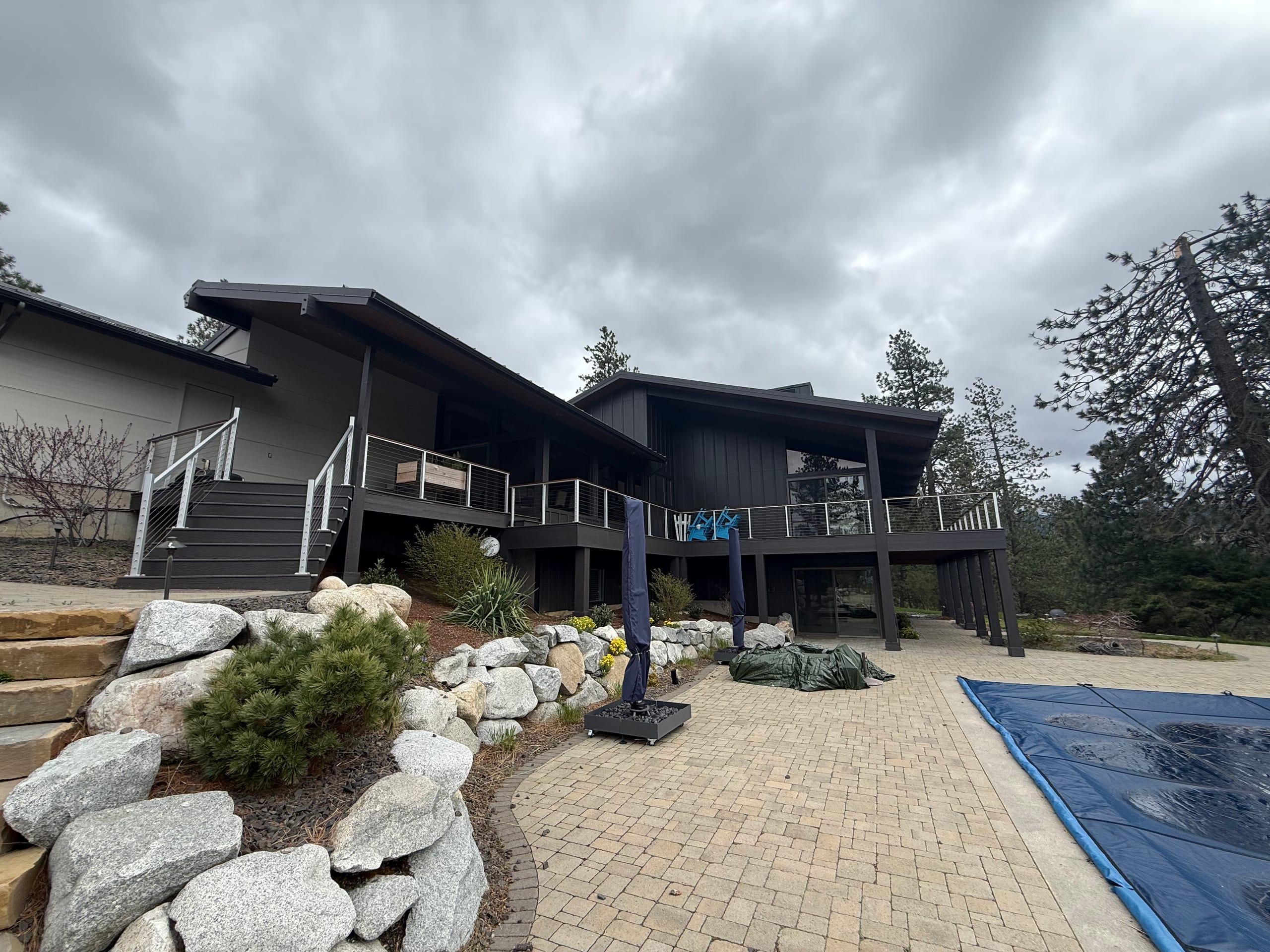 Dark contemporary home with elevated deck and stairs above a paver patio with pool cover and landscaped boulders, cloudy sky