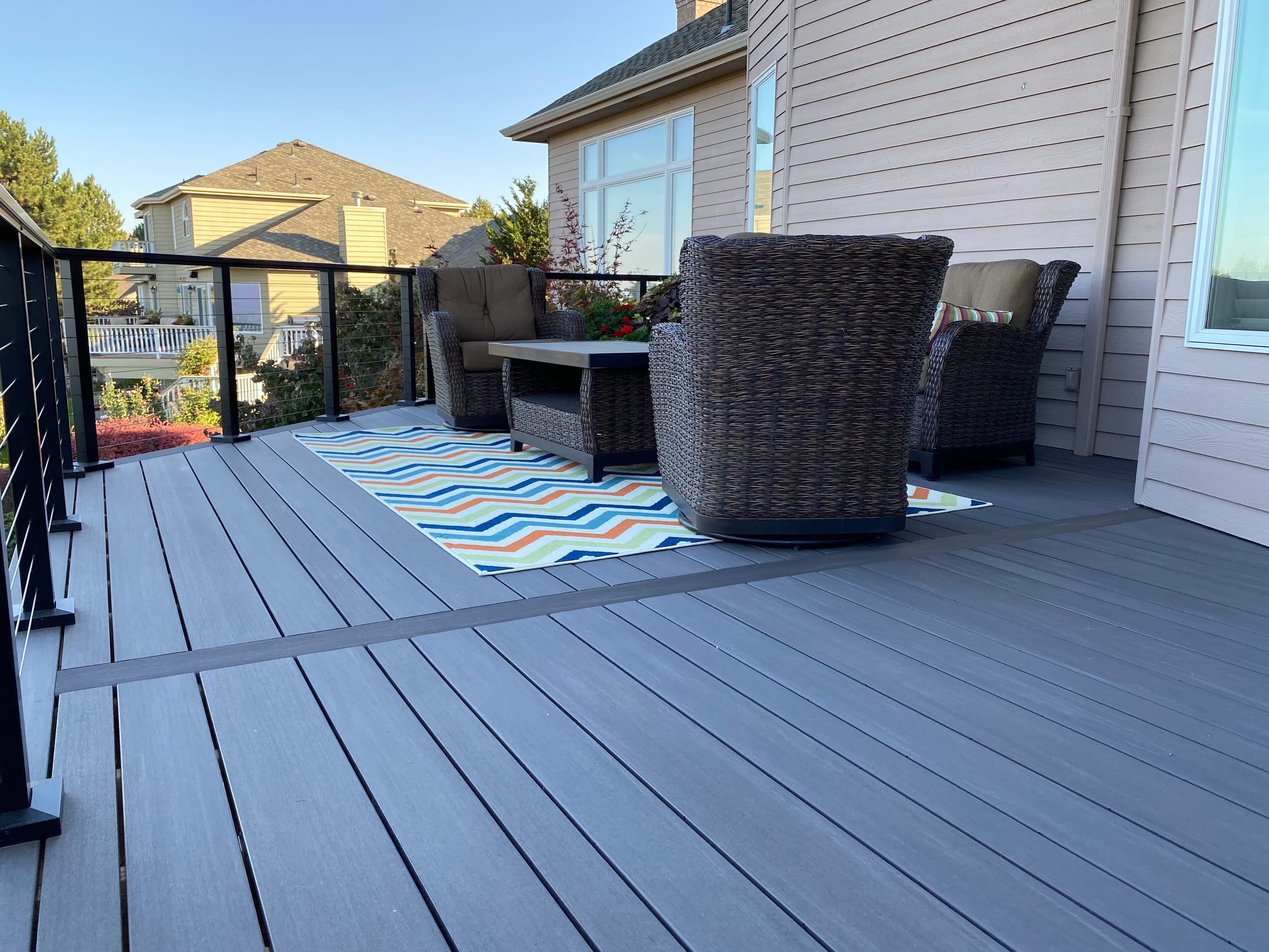 Dark grey composite deck with striped rug, wicker lounge furniture and black cable railing beside house