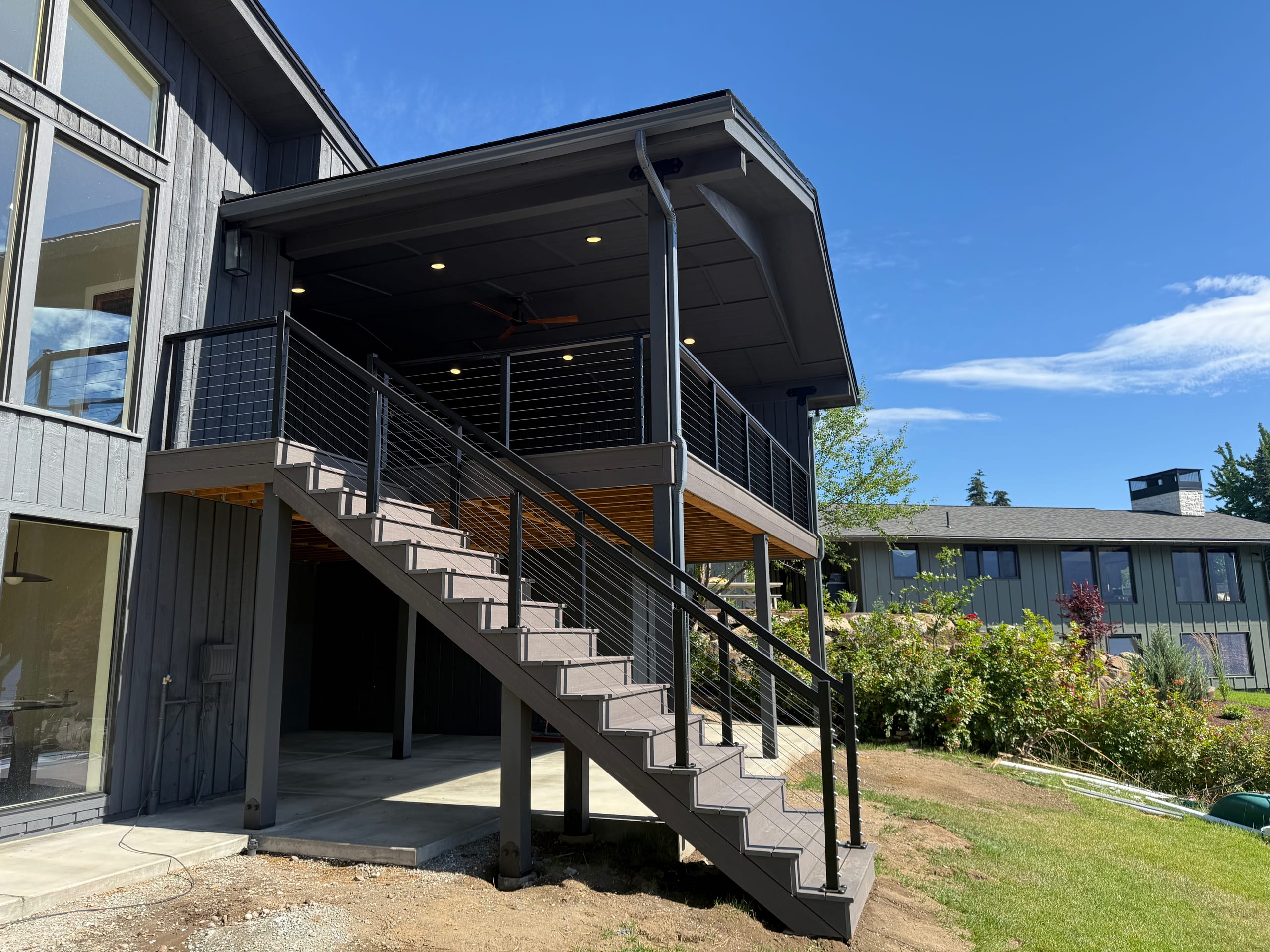Dark house with gabled covered deck, horizontal cable railing, wide composite stairs and posts over concrete patio, blue sky