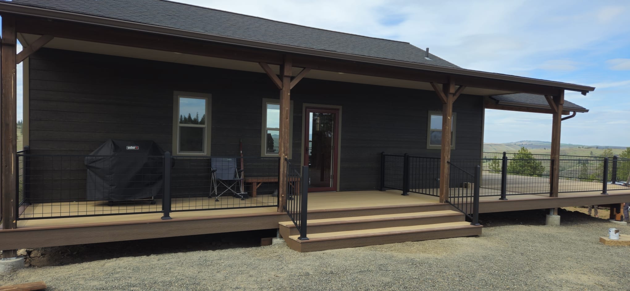 Dark house with long covered wraparound porch on brown deck with timber posts, black railing and prairie view