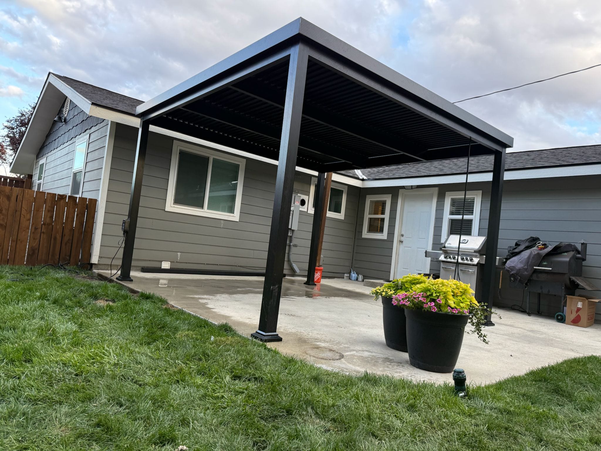 Dark metal free-standing patio cover over a concrete patio with grill and planters beside a grey house