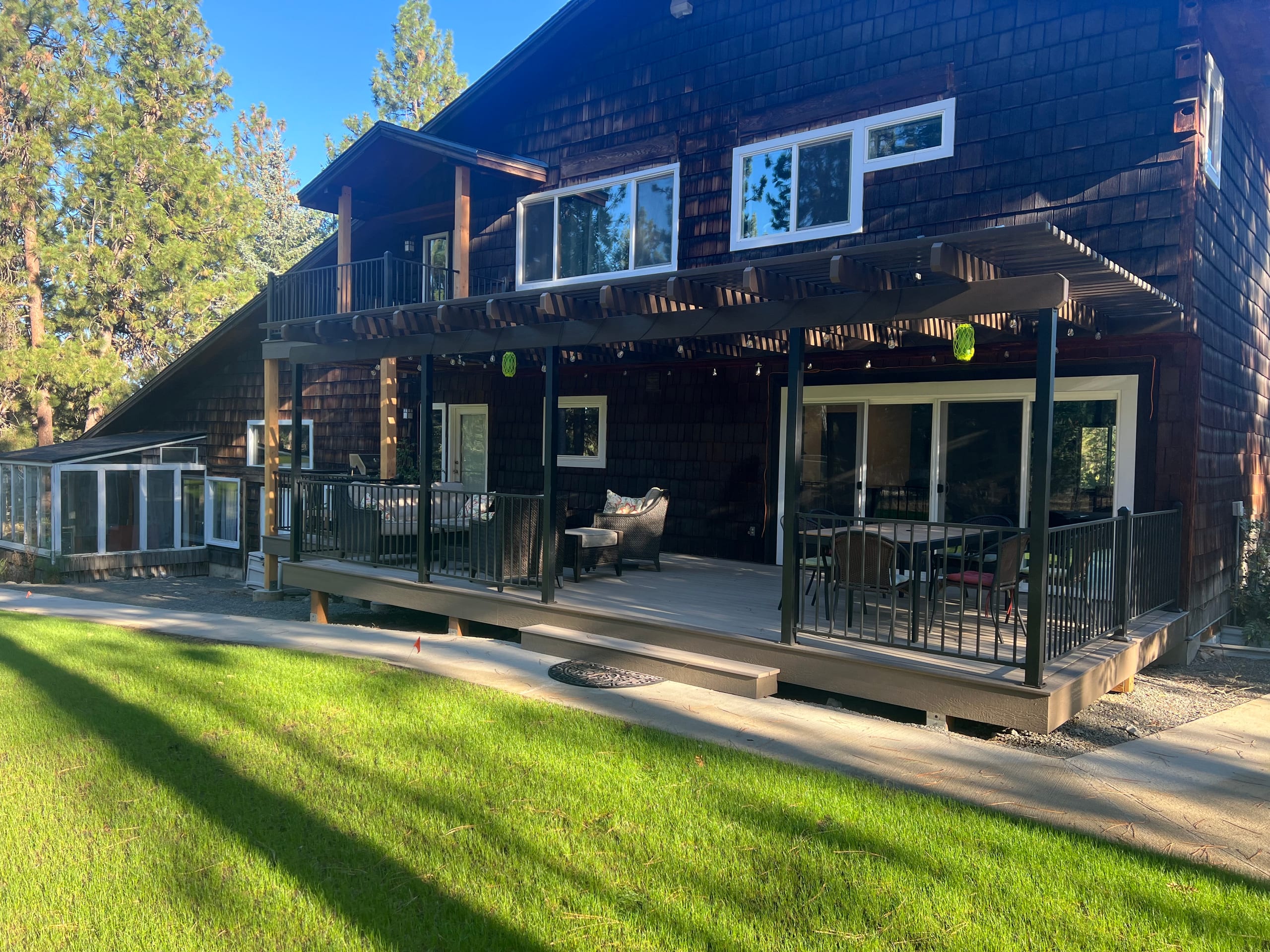 Dark shingle house with a tan composite deck, black railing and a pergola with cross slats above, lawn and pines
