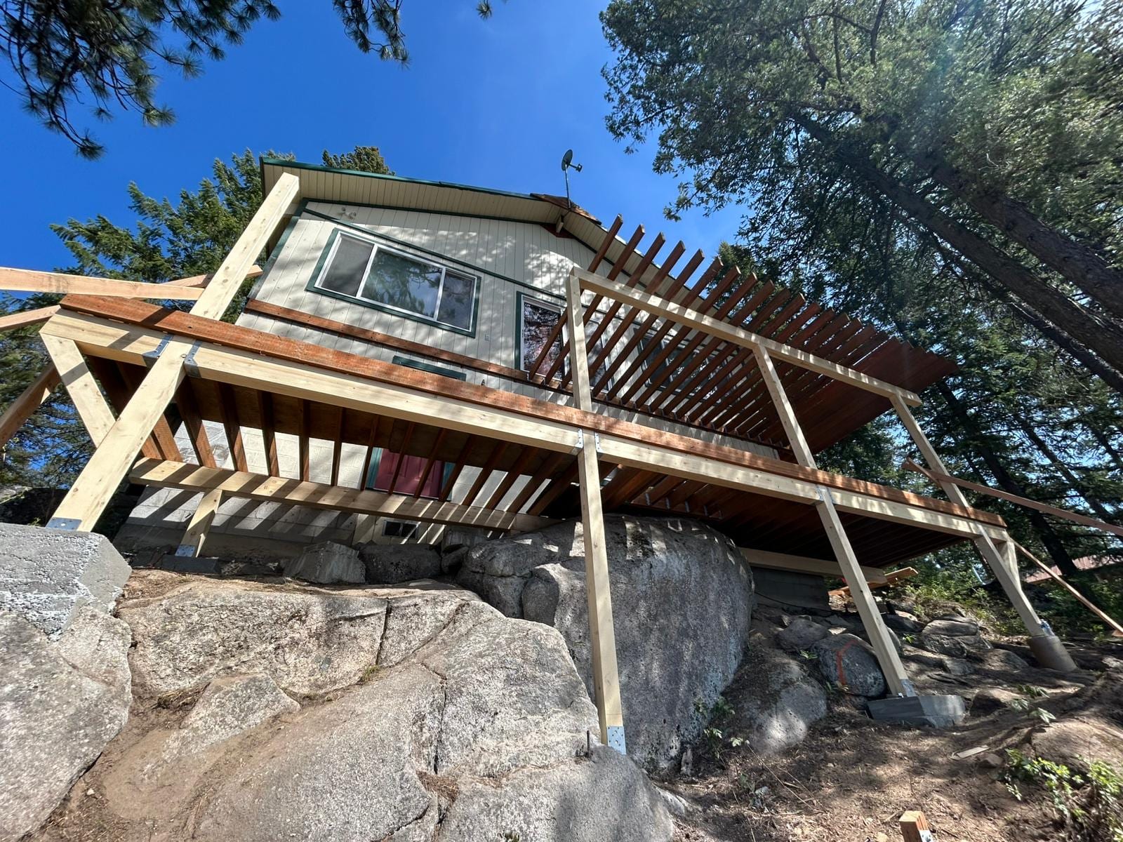 Deck with cedar pergola framing built on timber posts atop large granite boulders, seen from below, trees behind