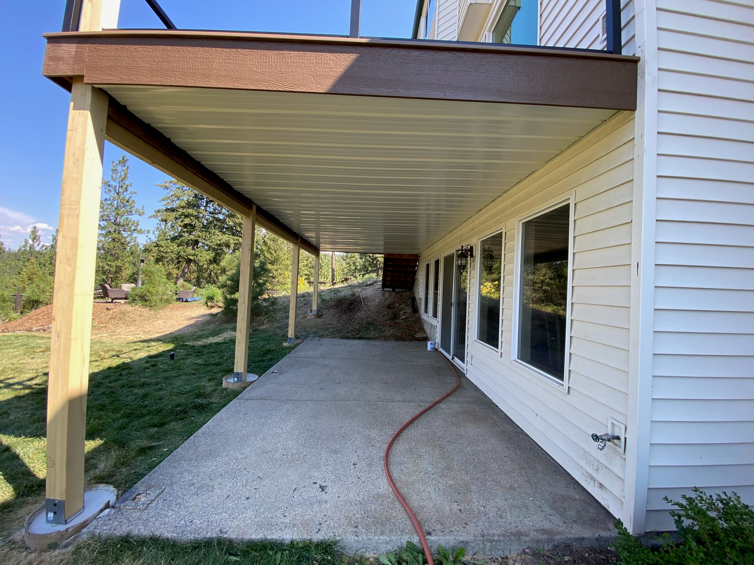 Deck underdeck system with white ceiling and wood posts above concrete patio along white siding house