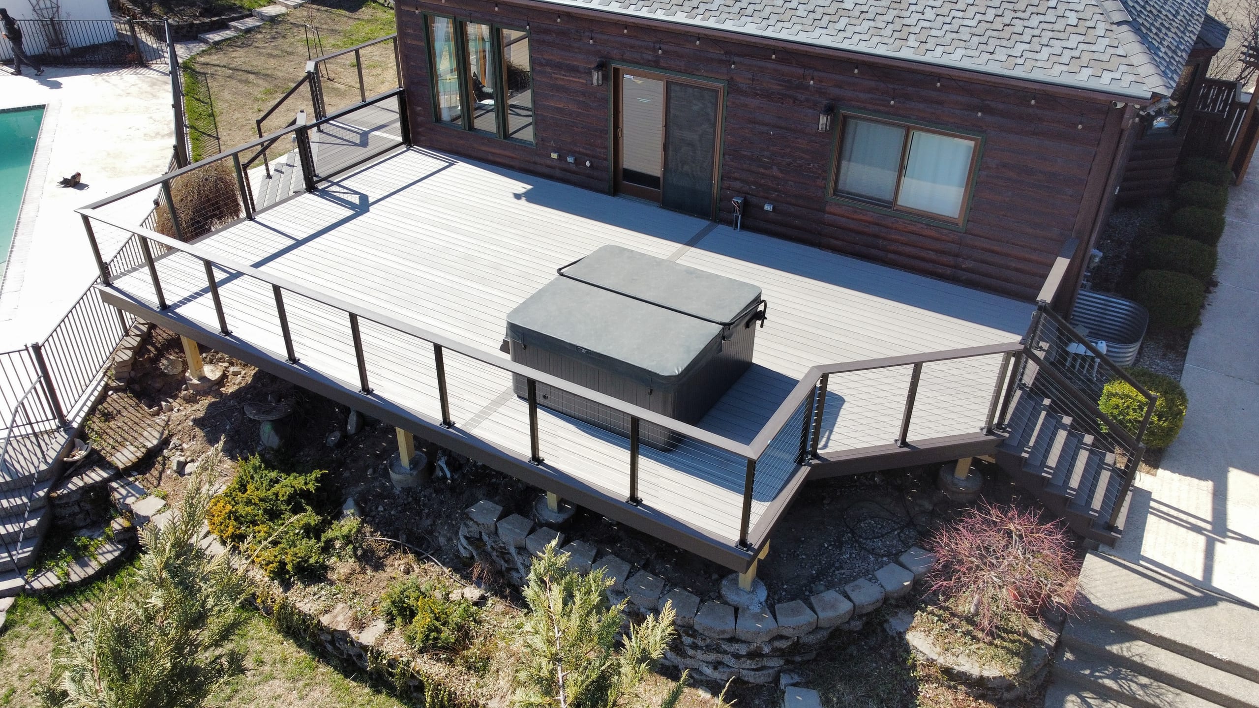 Drone view of grey composite deck with hot tub, cable-style railing and posts beside a pool and brown house