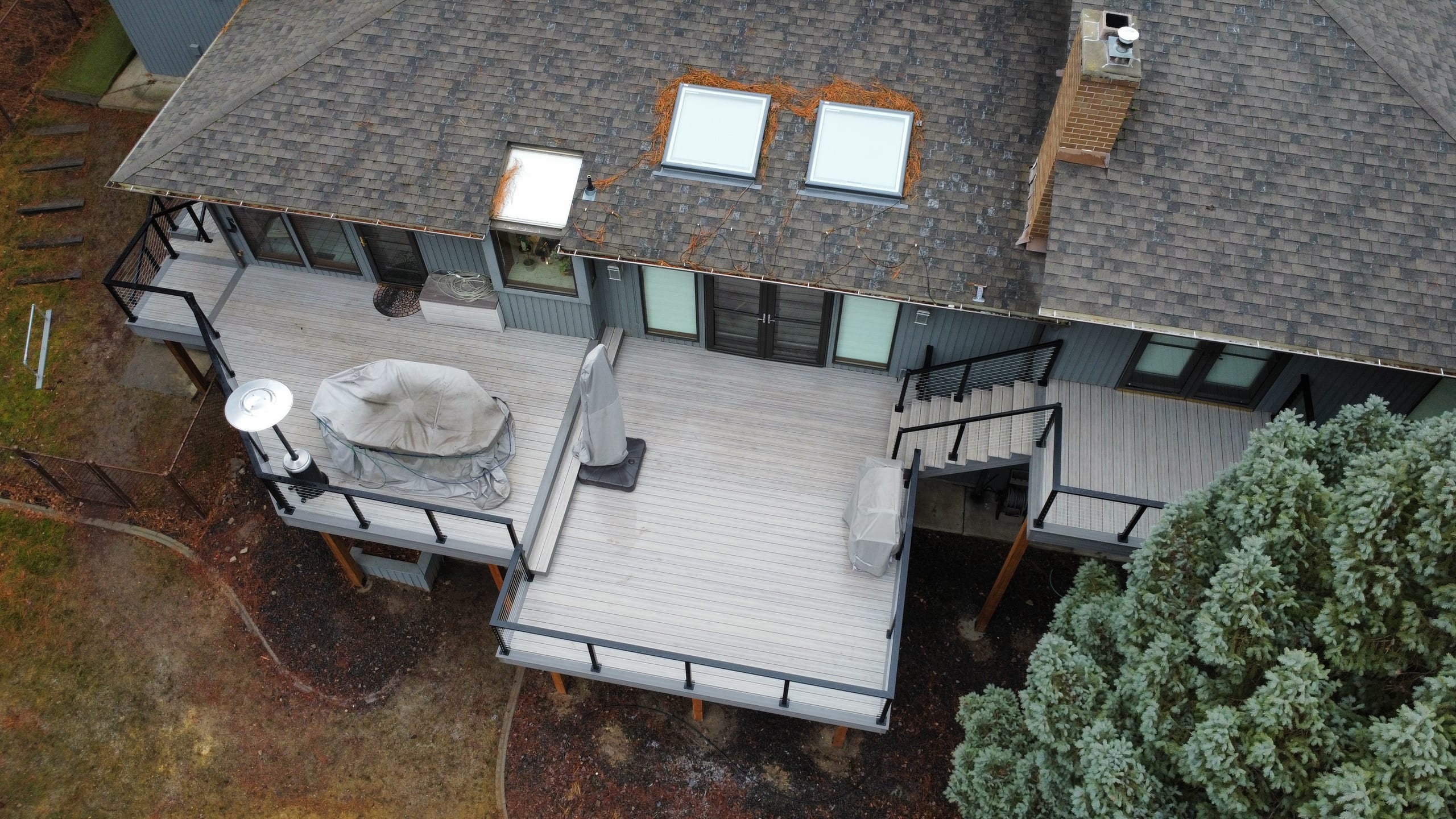 Drone view of light gray composite deck with black metal railing, covered hot tub and grill beside a gray cedar-trimmed home