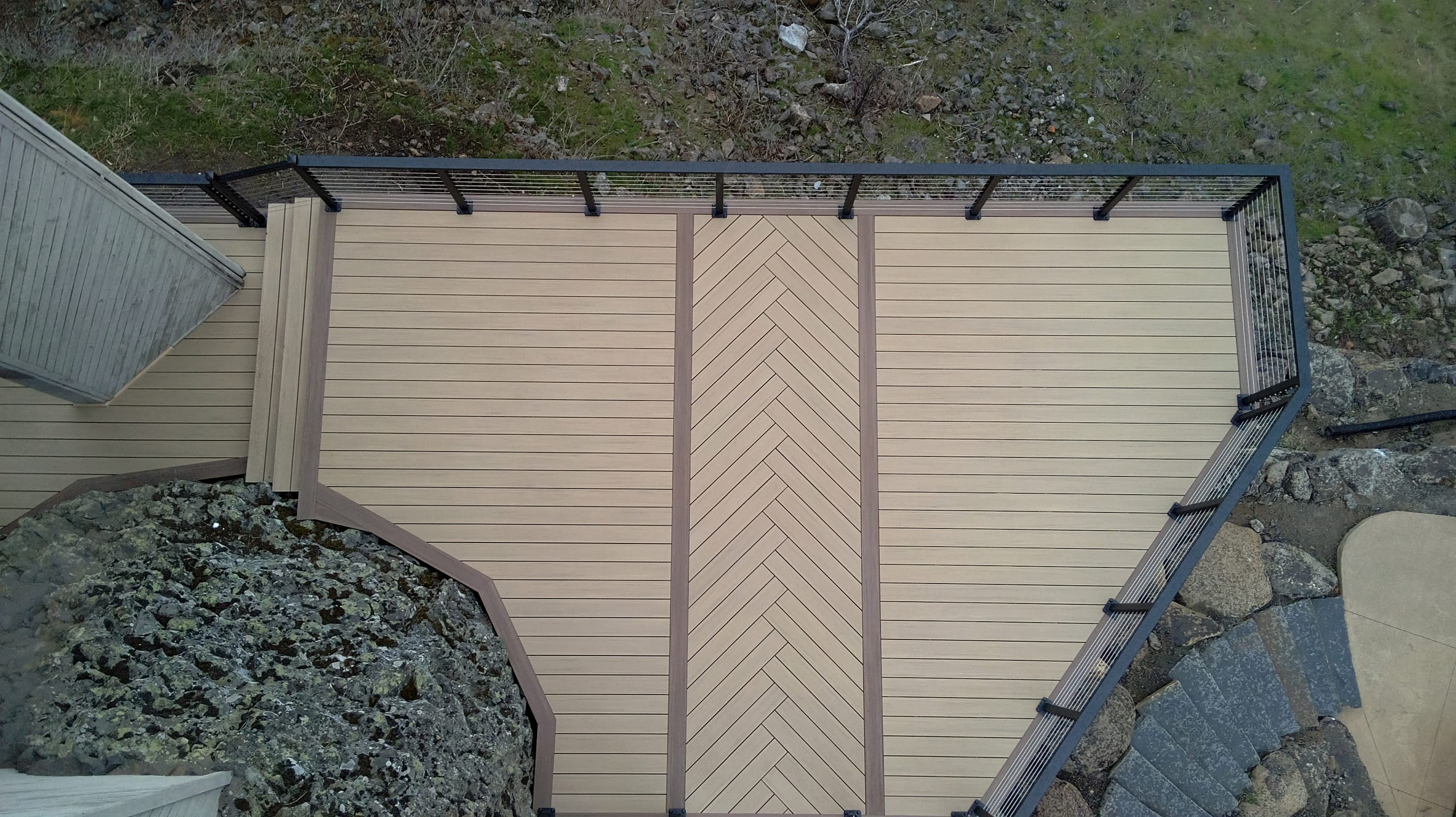 Drone top-down of tan composite deck with chevron center inlay, dark border and black railing beside a rock planter