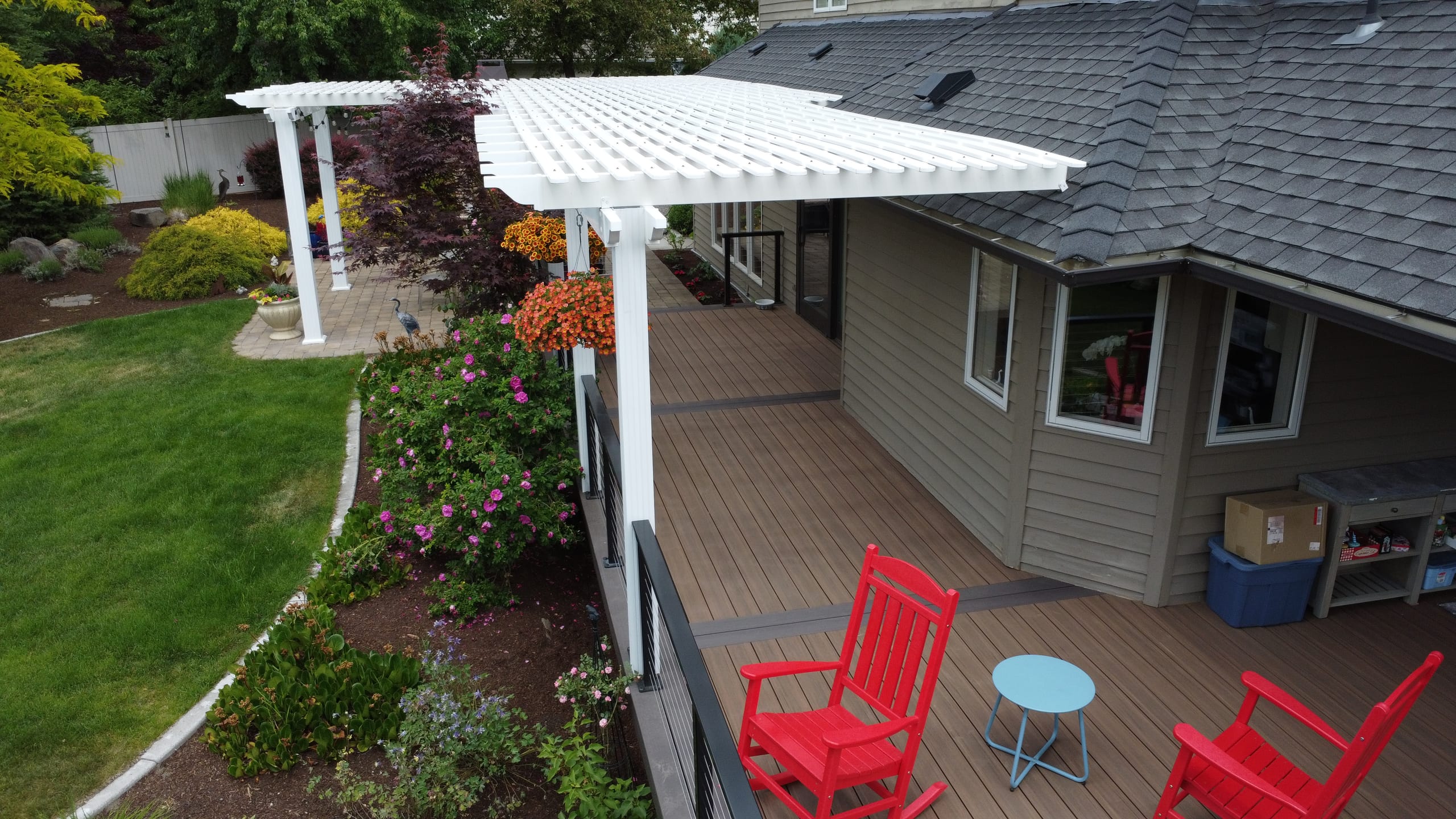 Drone view of white pergola over brown composite deck with red rocking chairs and lawn beside house