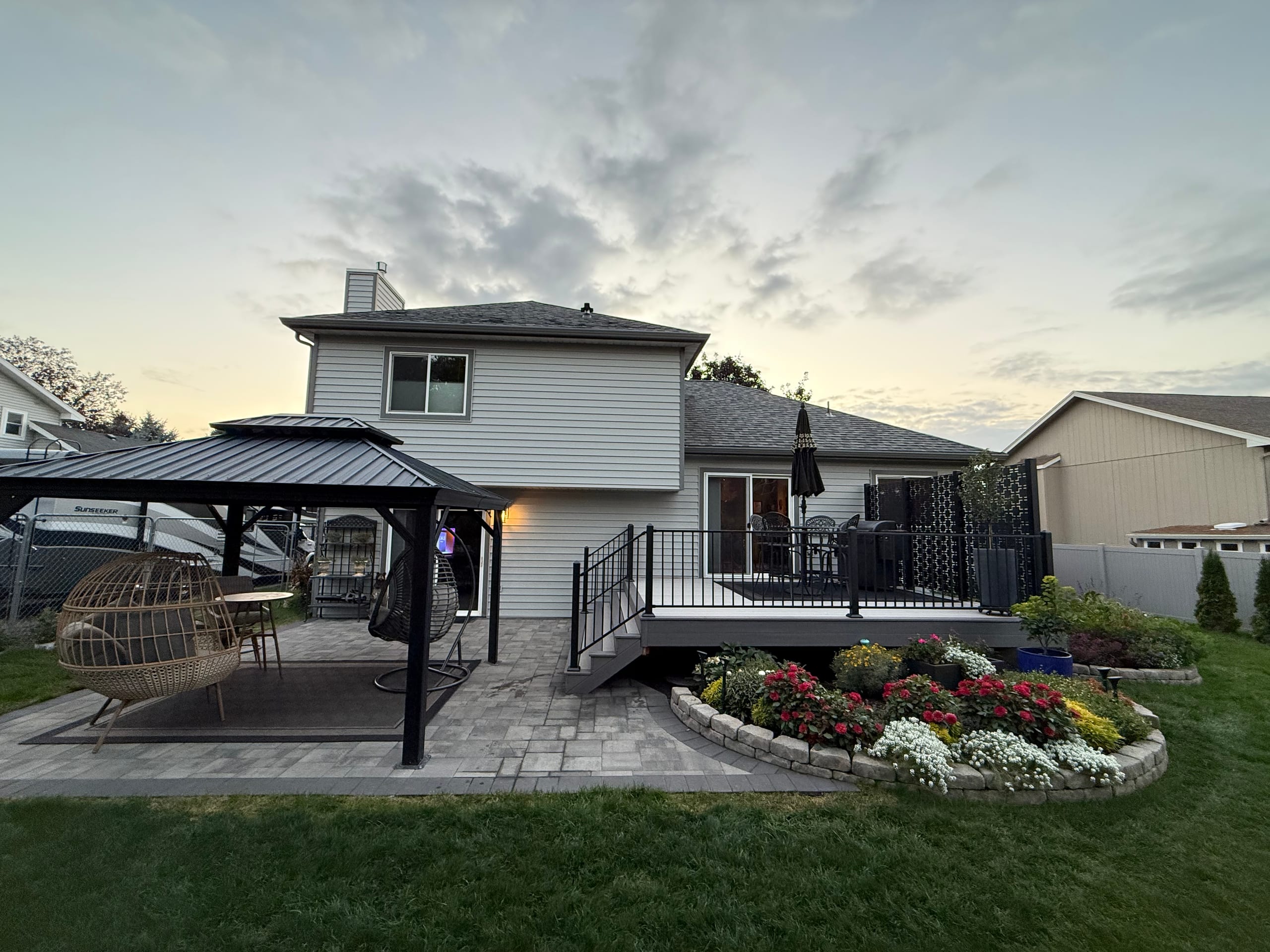 Dusk view of backyard with gray deck, black railing, paver patio, black gazebo, seating and flower bed