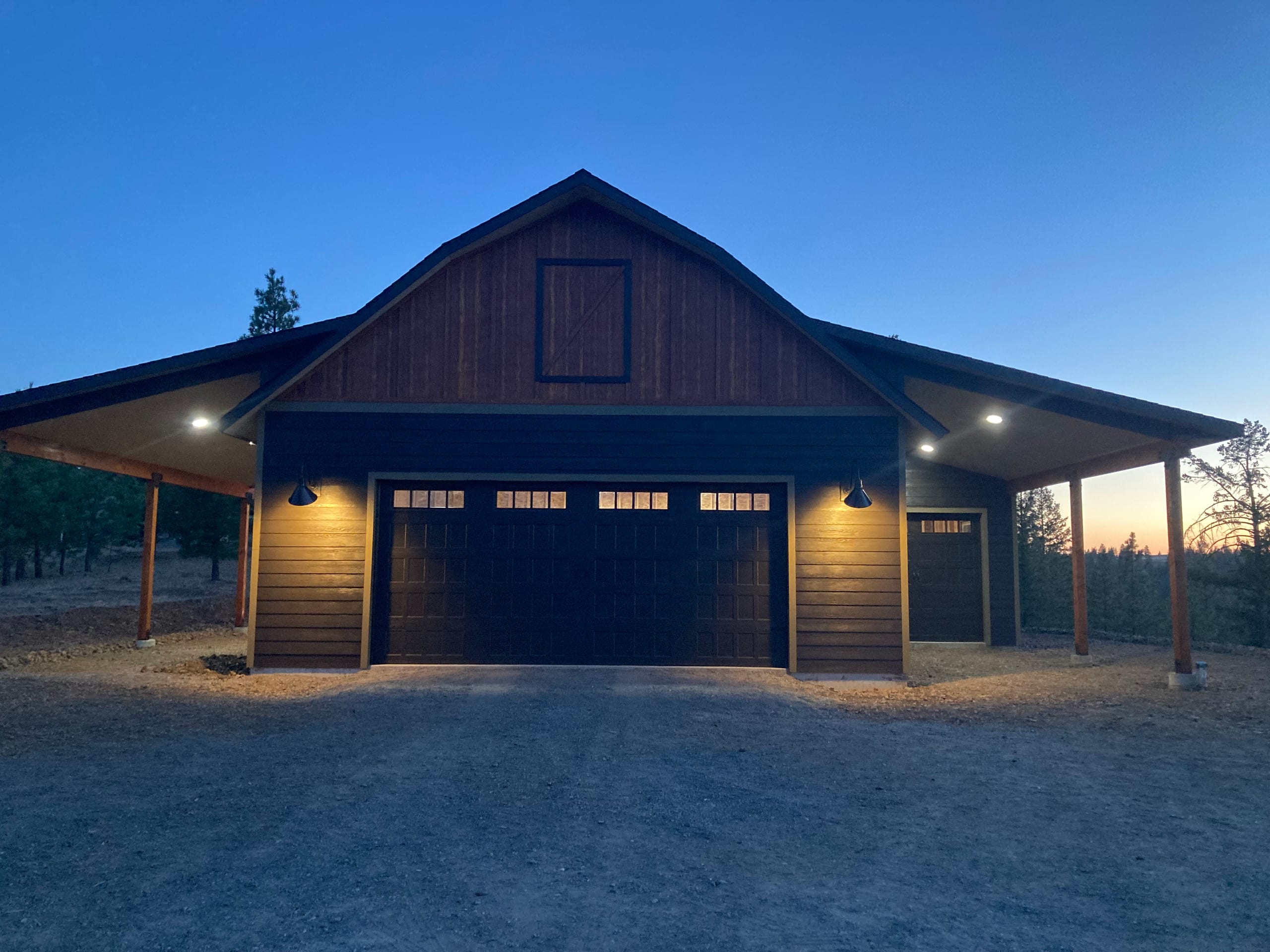 Dusk view of dark barn-style garage with lit carport soffits and wood-clad gable, gravel apron