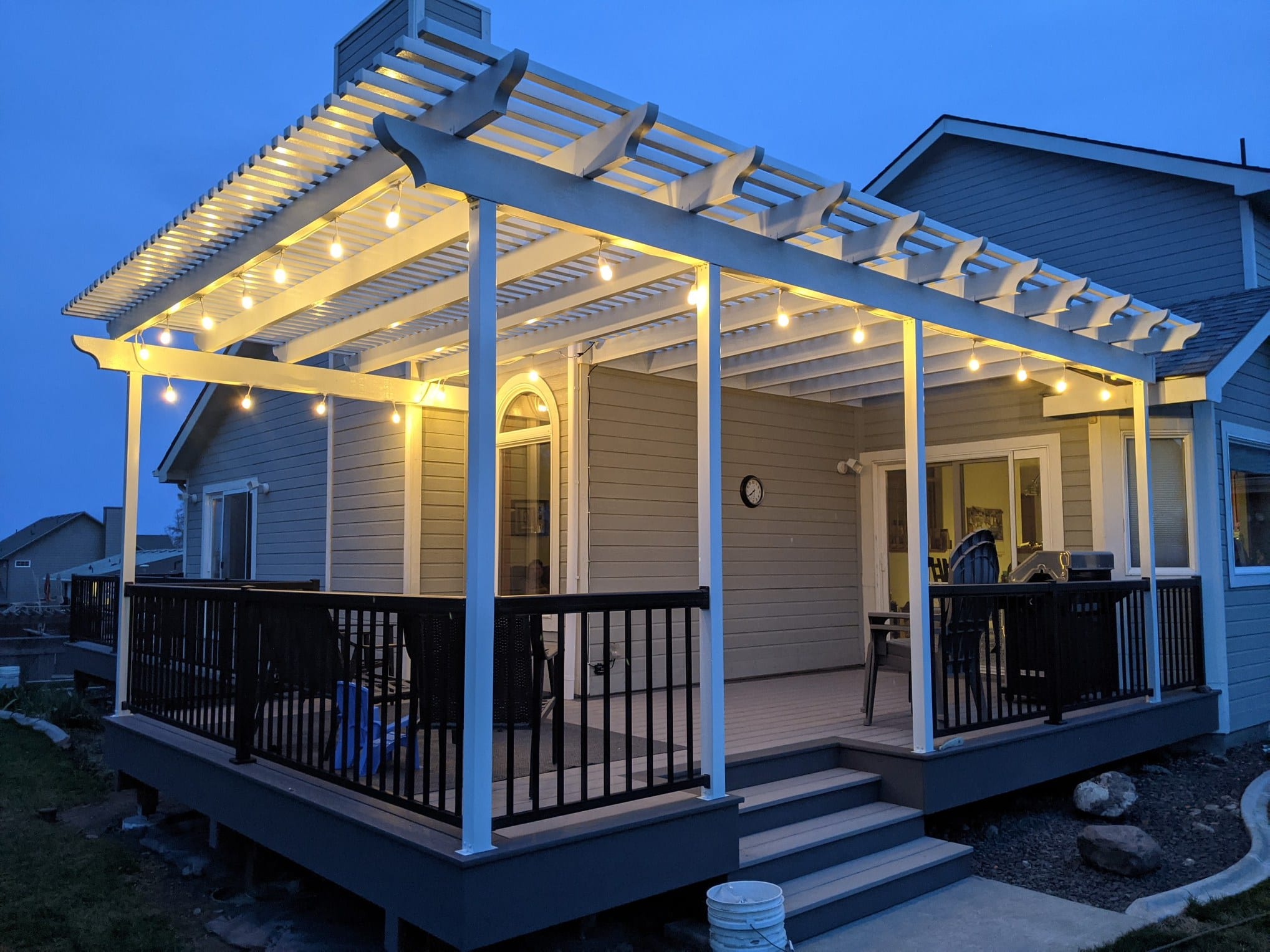 Dusk view of white pergola with string lights over a covered deck, black metal railing and steps to the yard