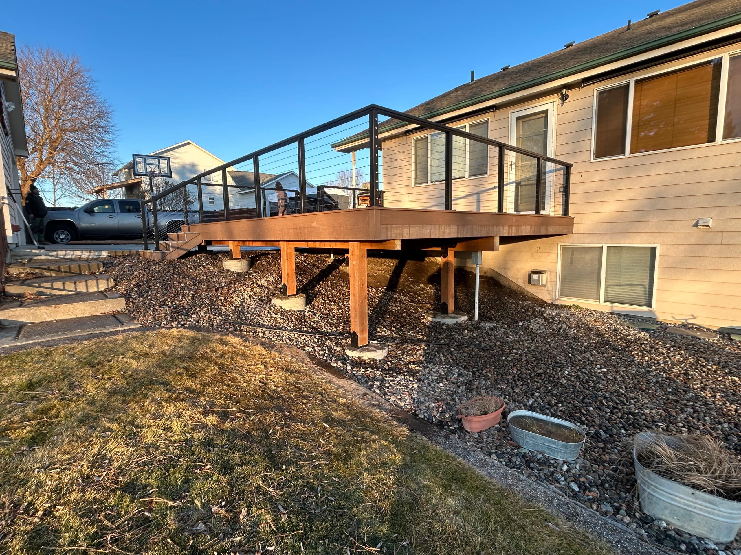 Elevated brown deck with cable railing on wood posts over gravel slope beside a tan house