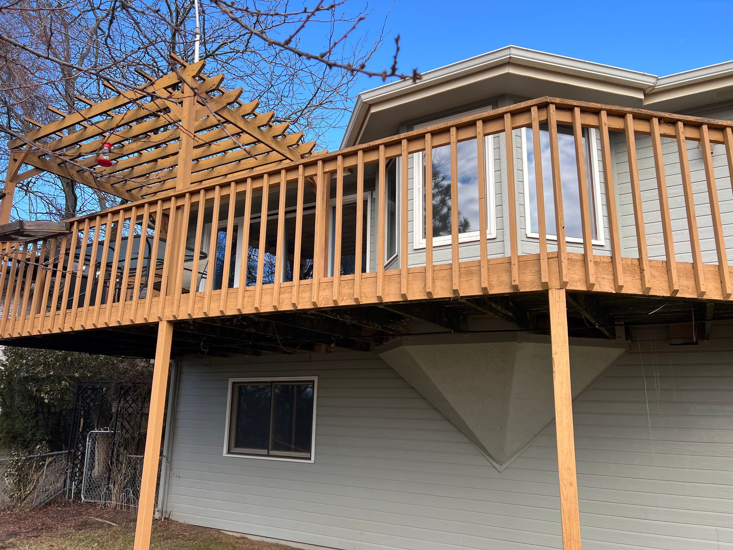Elevated cedar-stained wood deck with wood picket railing and pergola corner on grey house, blue sky