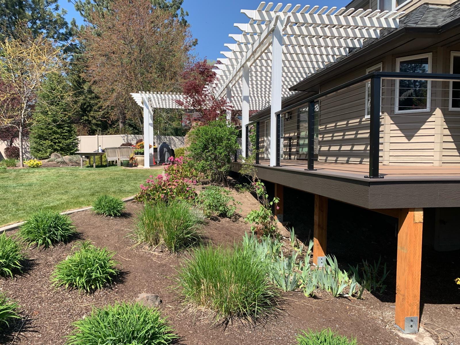 Elevated composite deck with cable railing and white pergola along the house, sloped landscaped garden below