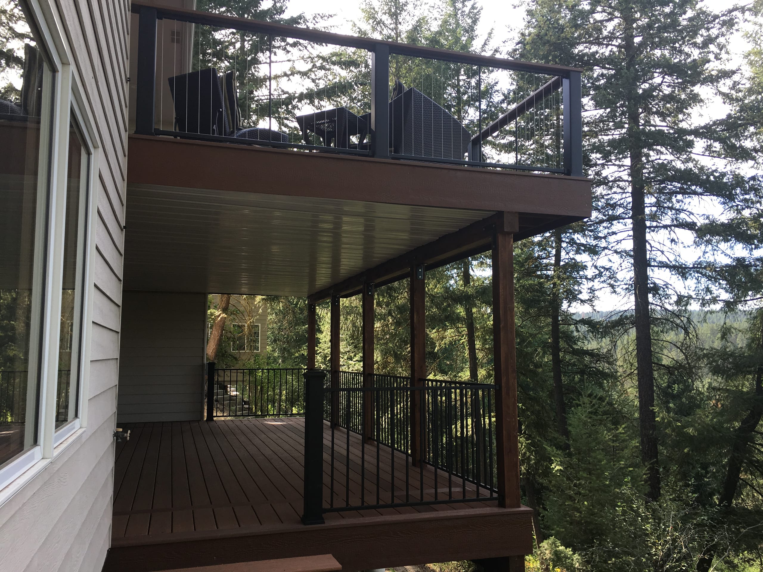 Elevated dark composite deck with black metal picket railing over a covered lower patio, surrounded by pine trees
