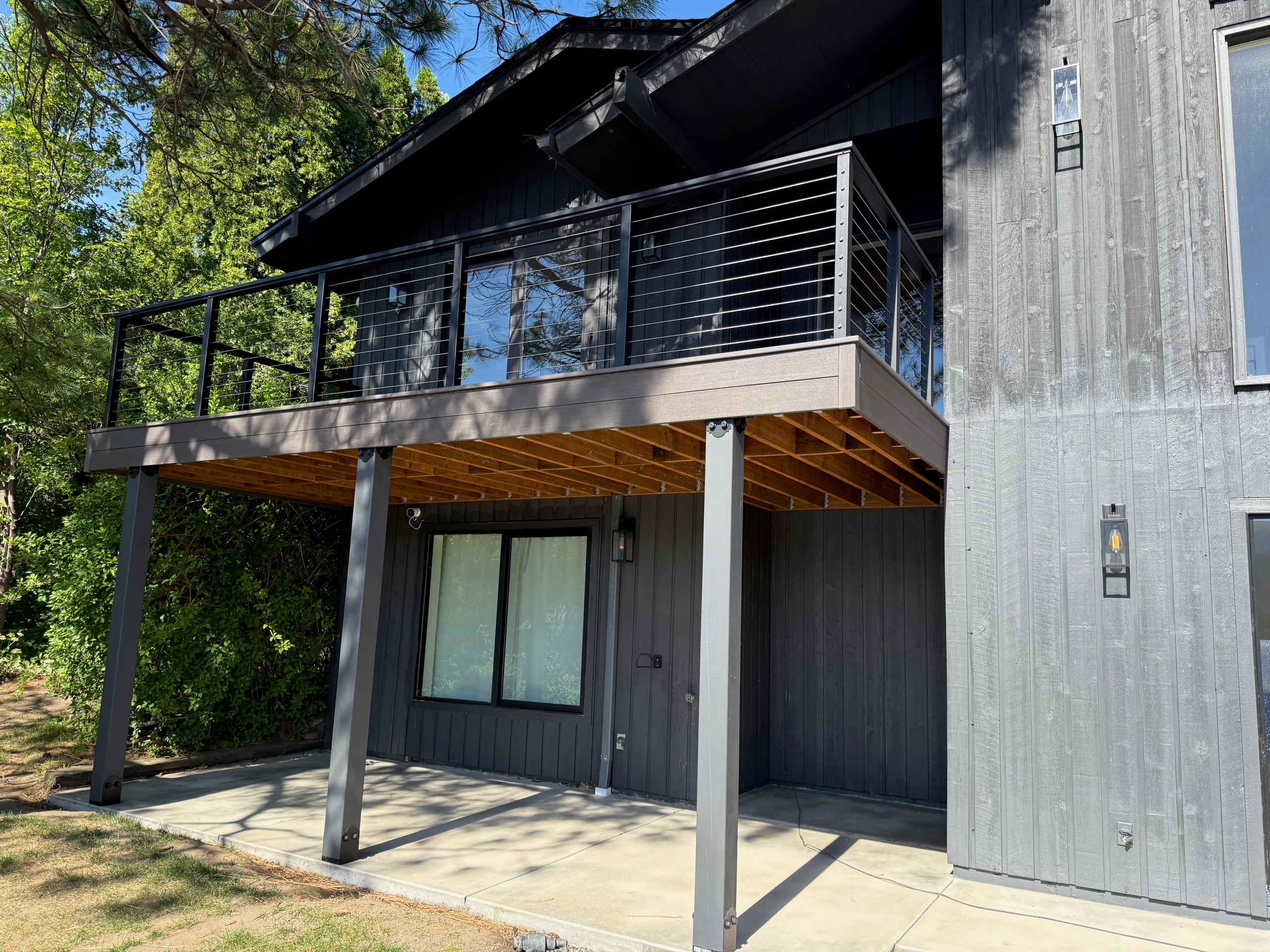 Elevated dark composite deck with cable railing on black posts, cedar-stained joists underneath, dark vertical-siding house
