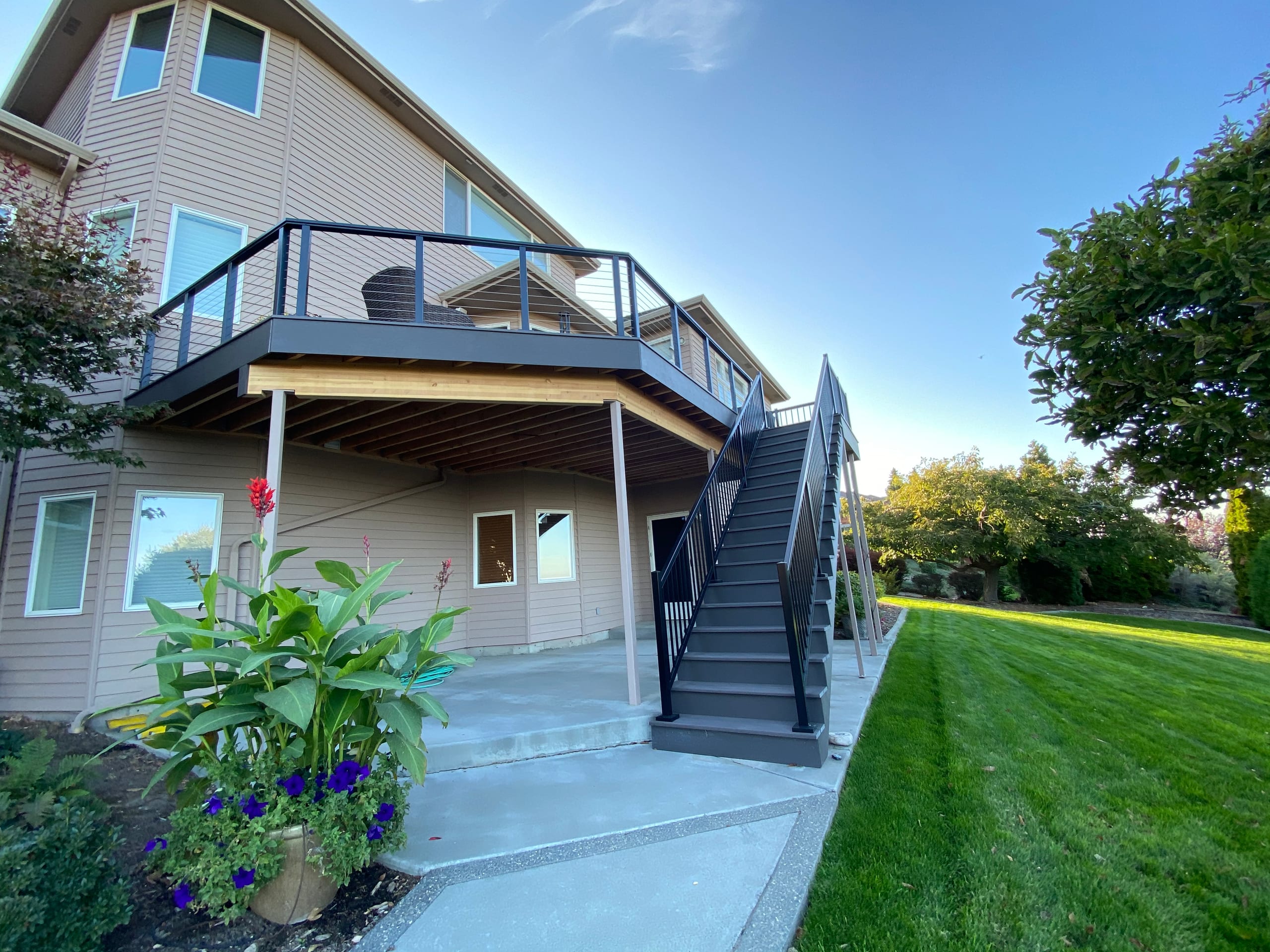 Elevated dark deck with cable railing and stairs over a covered concrete patio below, evening light
