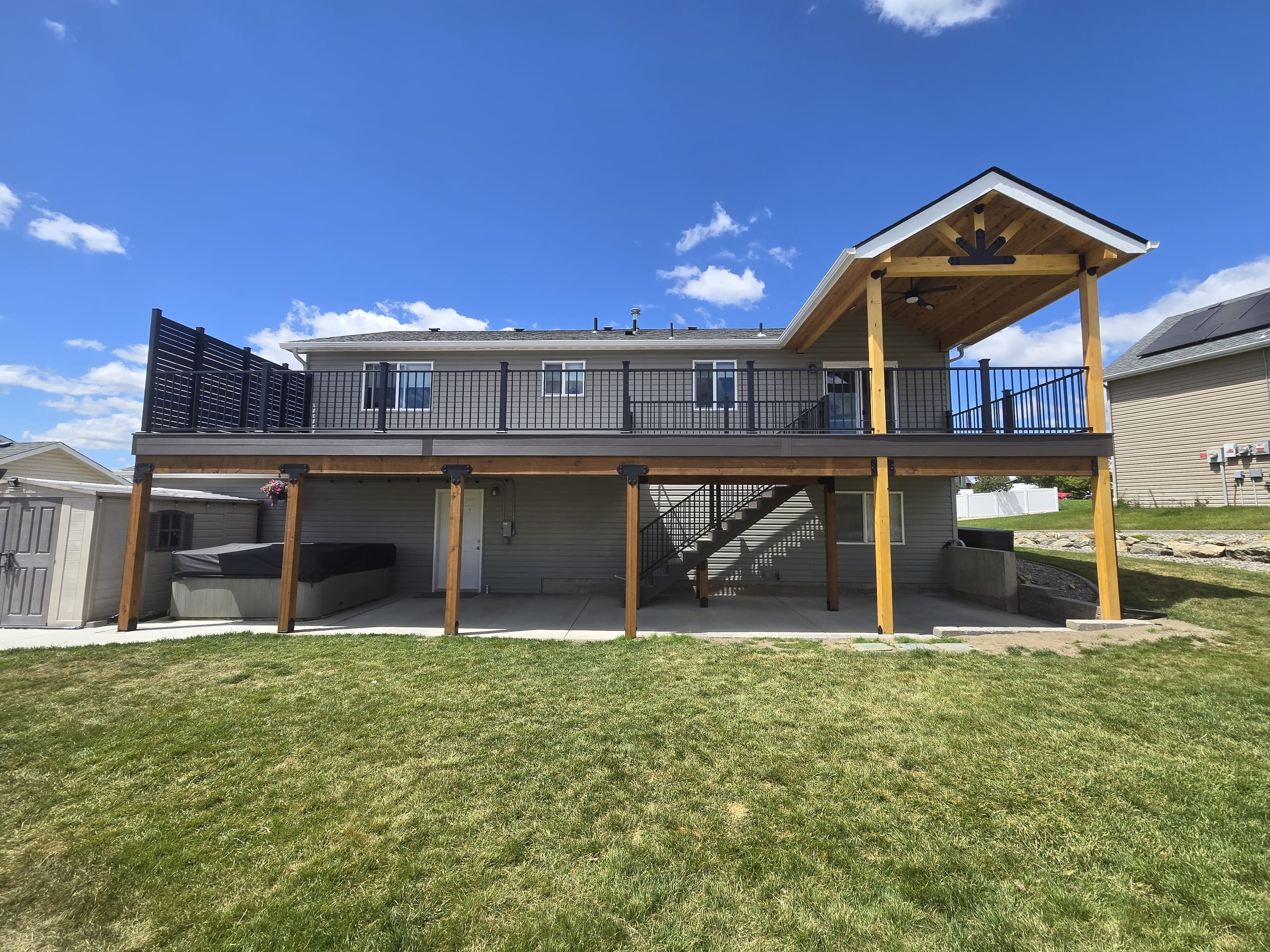 Elevated deck across a two-story house with gable-roof section, black railing, stained posts and patio beneath, green lawn