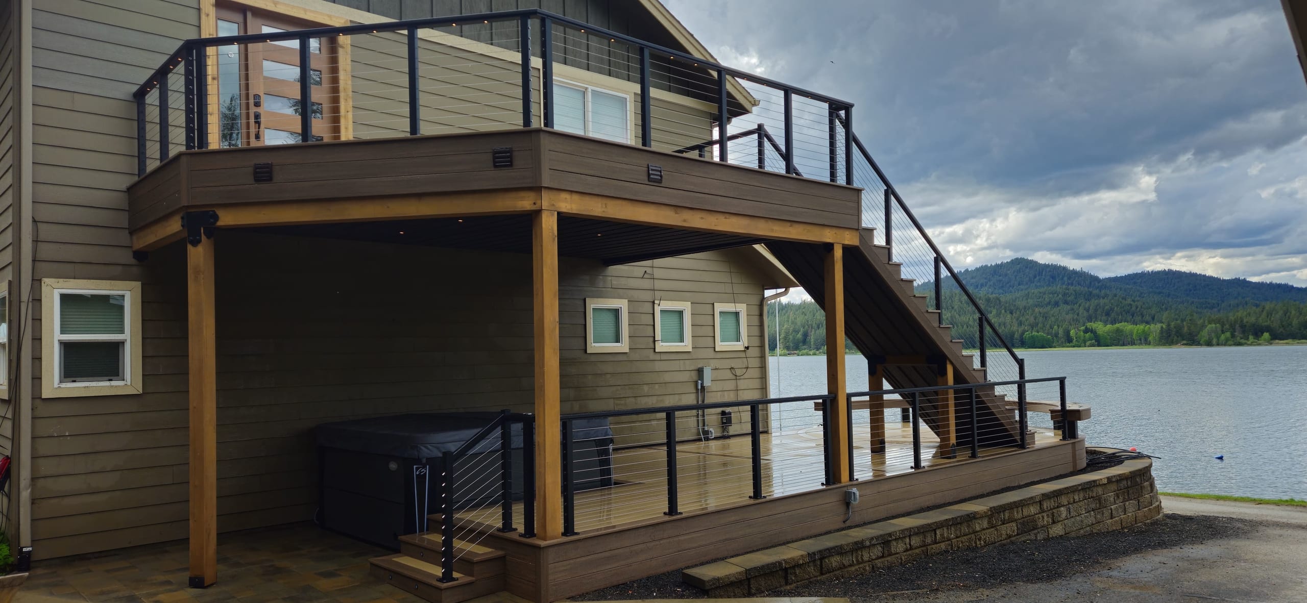 Elevated deck with black cable-style railing and stairs on cedar posts, hot tub below, lake and mountains behind