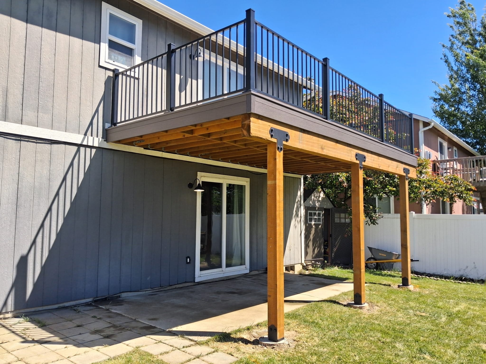 Elevated deck with black railing on cedar posts over a paver patio at a blue-gray house, lawn and white fence