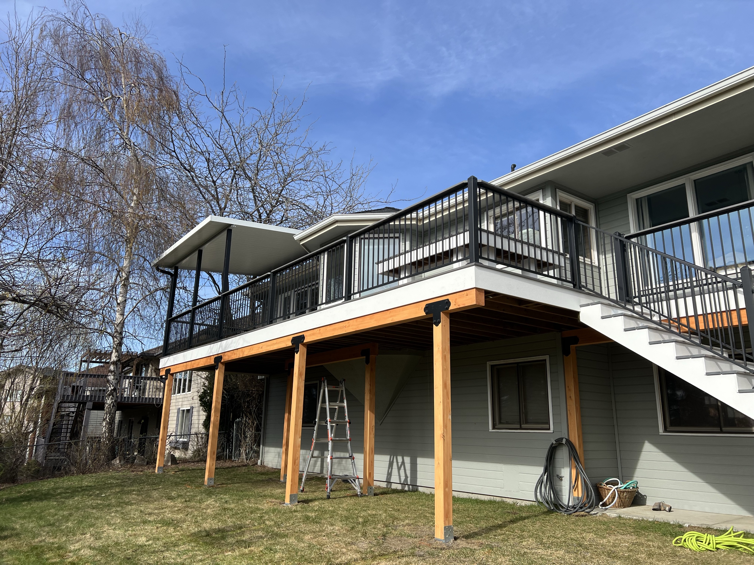 Elevated deck with black railing on cedar posts, white stair and underdeck on grey house, ladder and hoses on lawn
